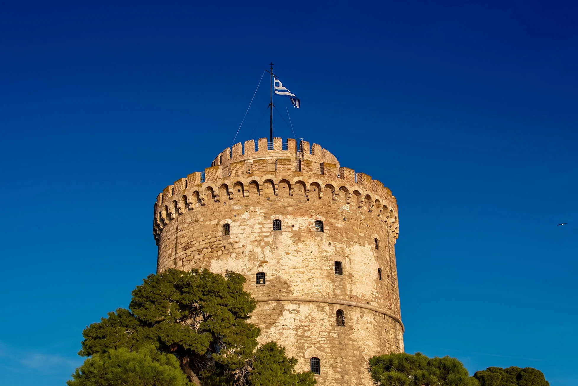 Ancient stone fortress tower with narrow windows, topped with a Greek flag, surrounded by green trees against a clear blue sky.