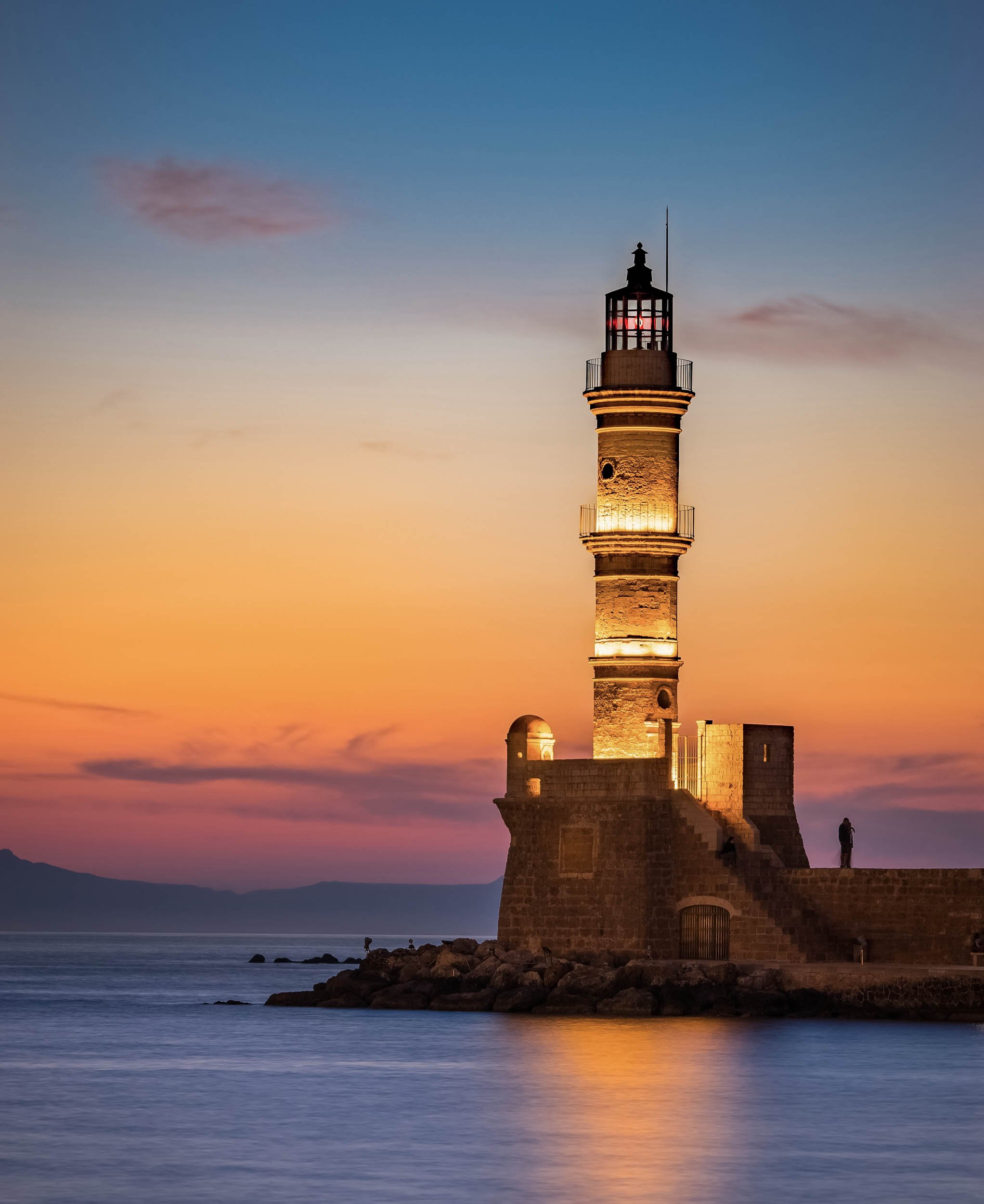 A lighthouse on a rocky shore during sunset with a person standing near the edge.