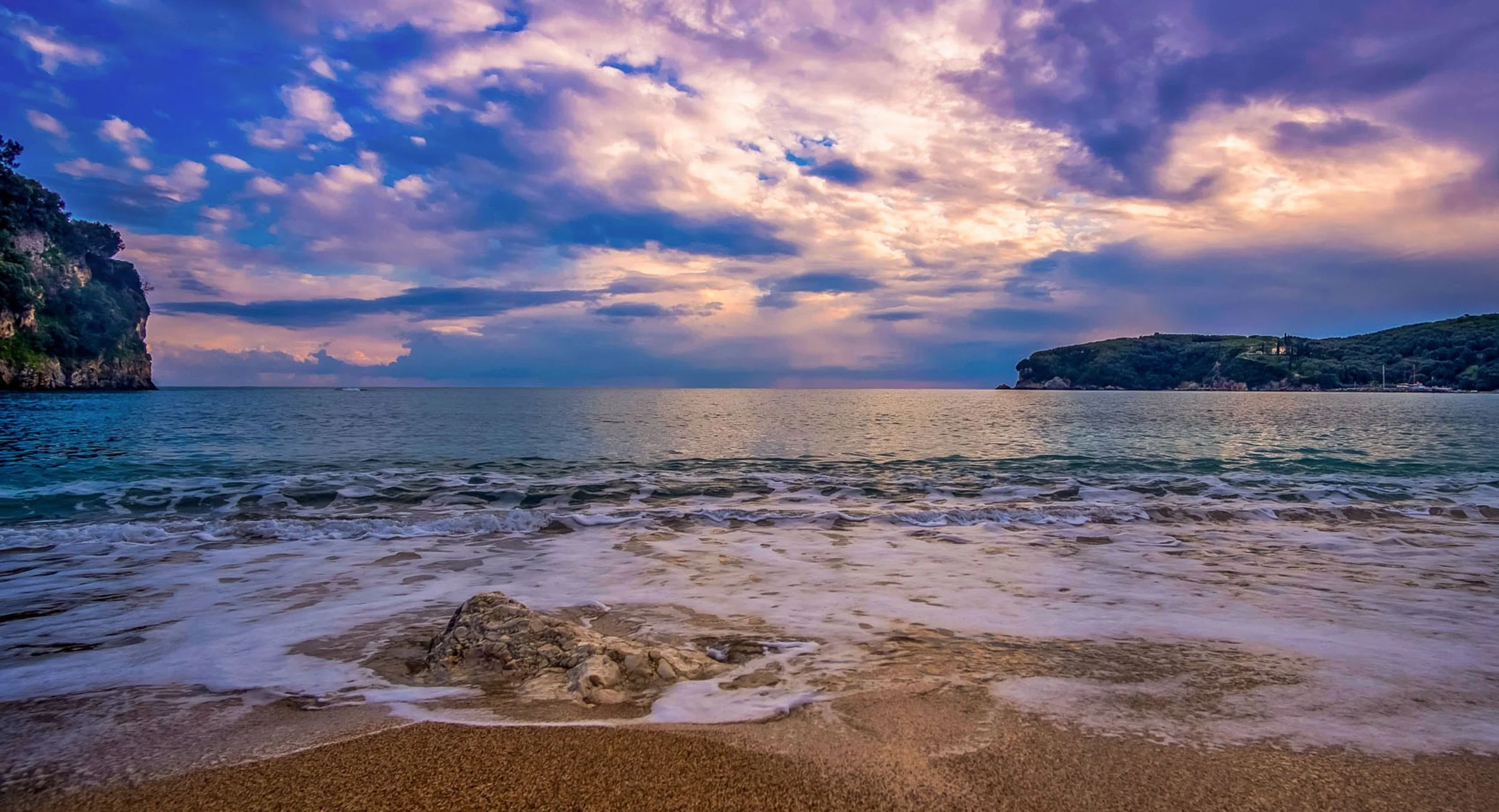 Scenic view of a beach with sand, gentle waves, and a rocky shoreline in the foreground, with hills and trees on either side under a partly cloudy sky during sunset or sunrise.