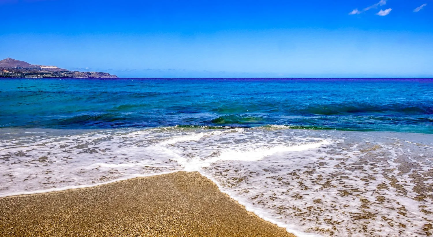 Calm ocean waves washing up on a sandy beach with cliffs in the distance under a blue sky with some clouds.