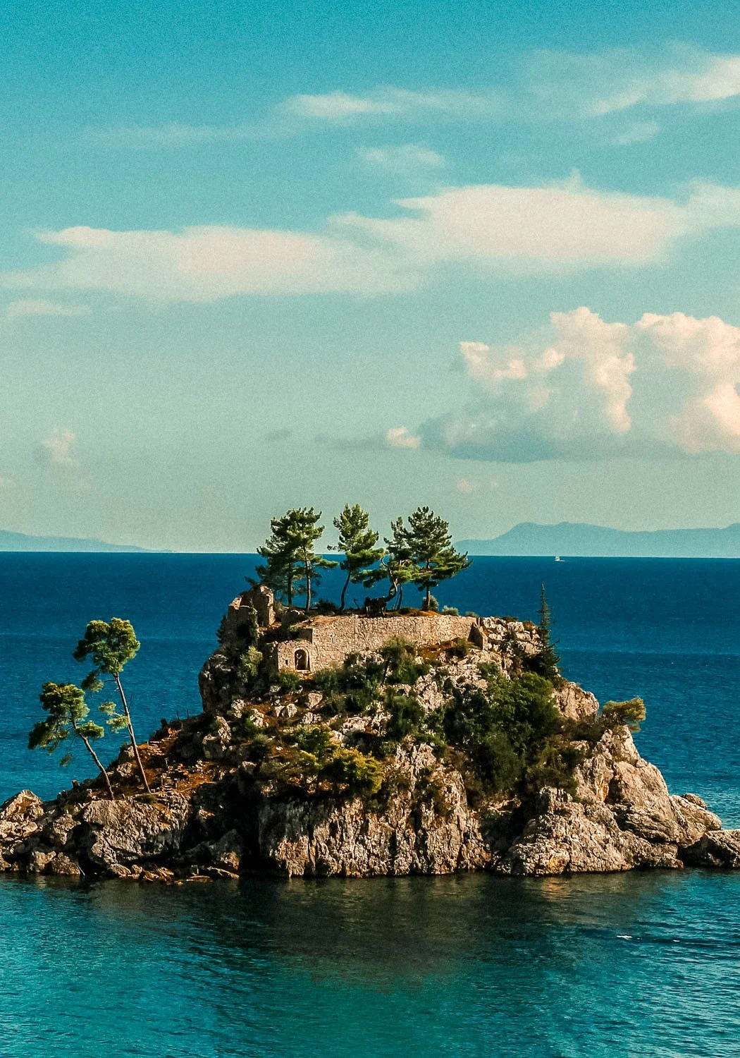 A small rocky island with trees and a stone structure, surrounded by blue water with a cloudy sky overhead.