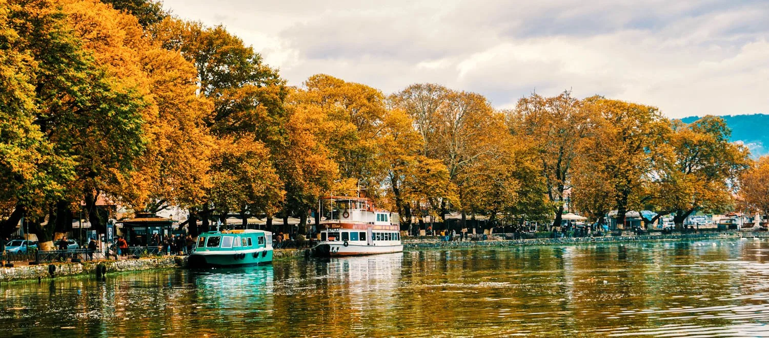 Boats docked by colorful fall trees along a lakeside or river with people walking along the waterfront.