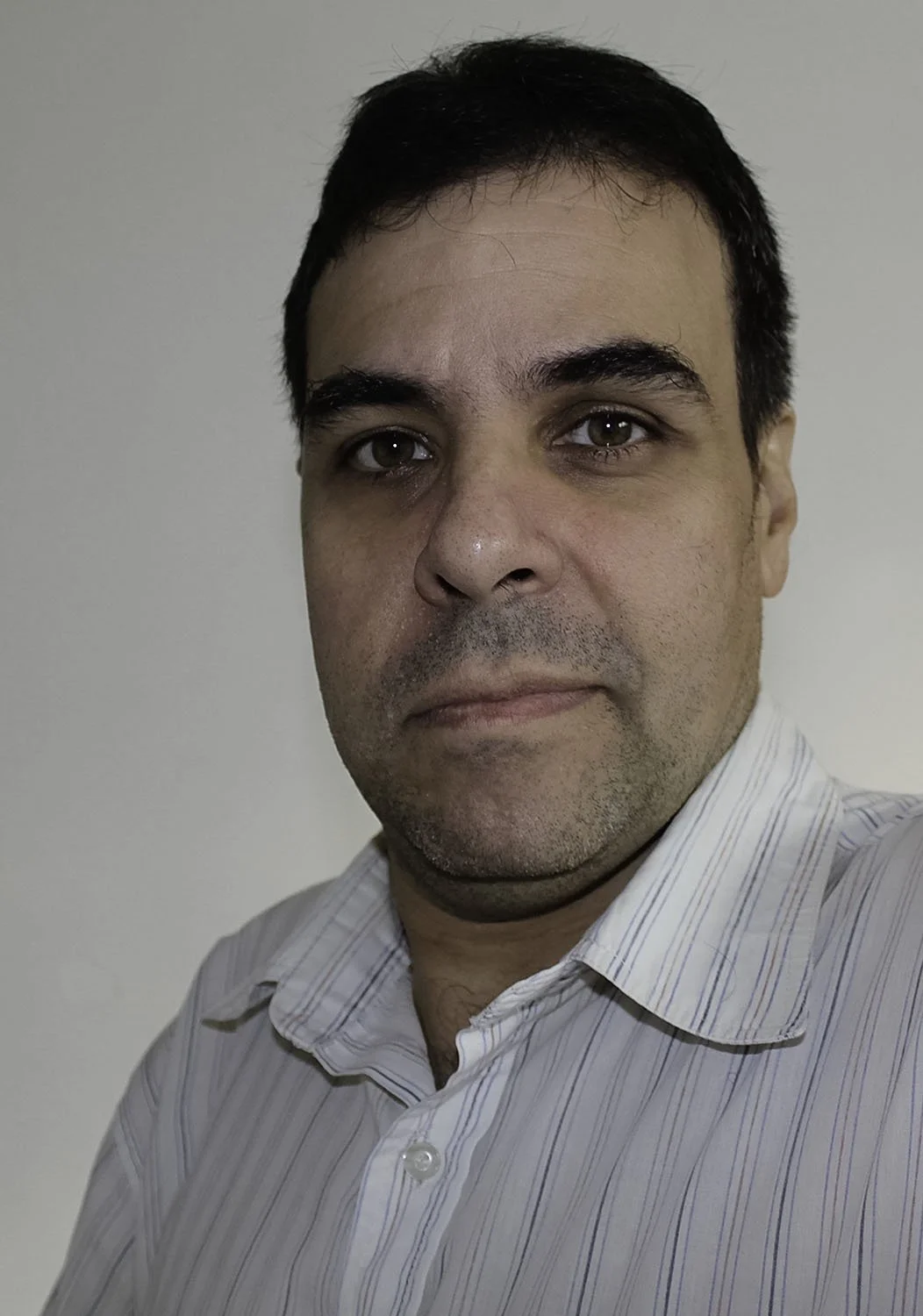 A man with short dark hair, wearing a light-colored, collared, striped shirt, looking at the camera with a neutral expression against a plain, light background.