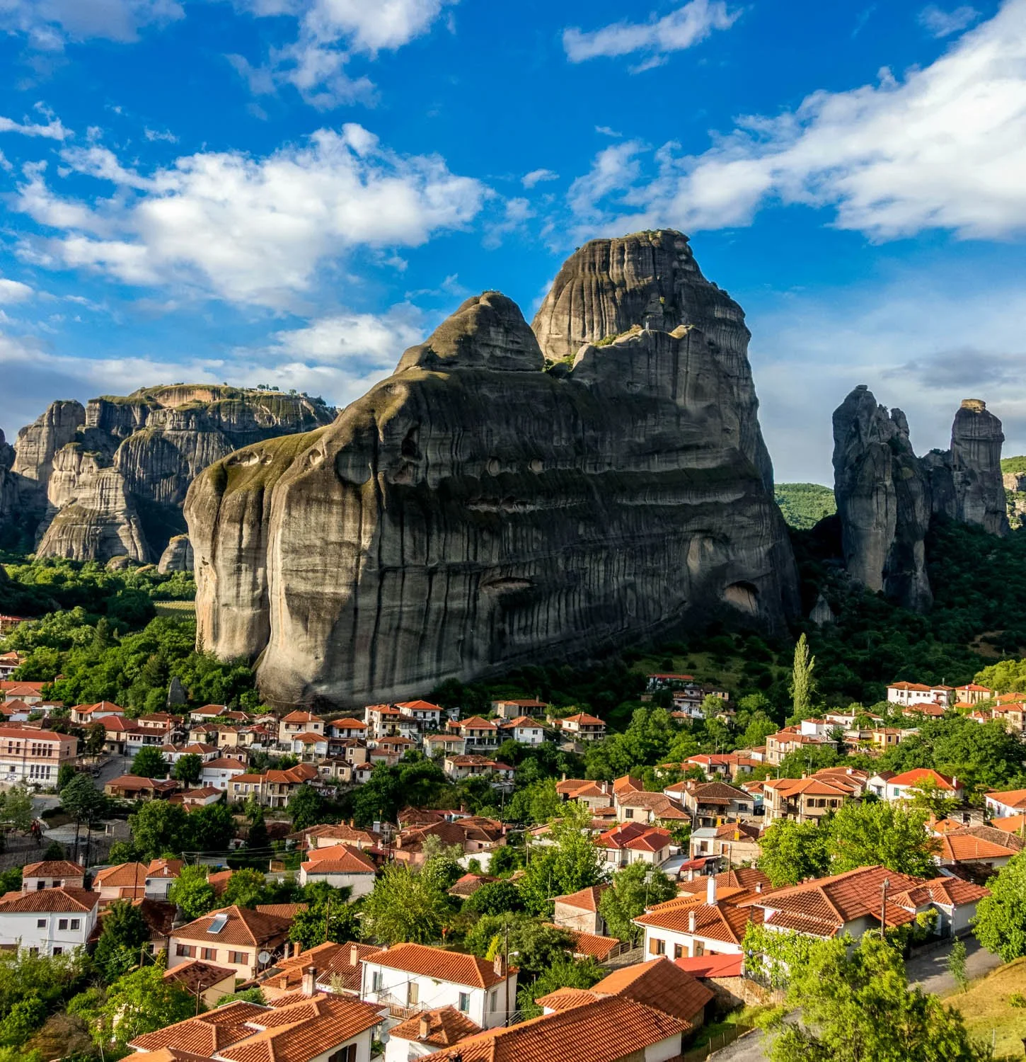 Village with red rooftops and green trees in foreground, large rocky mountains with vertical striations in midground, blue sky with white clouds in background.