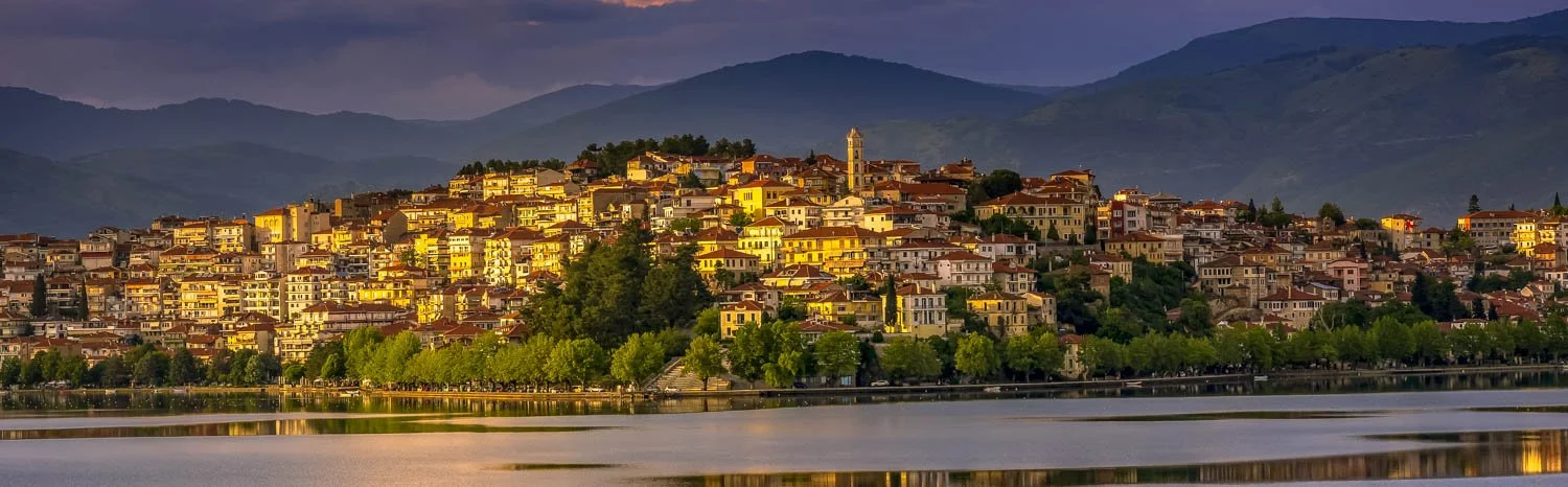A hillside Italian town with colorful buildings overlooking a river at sunset, with mountains in the background.
