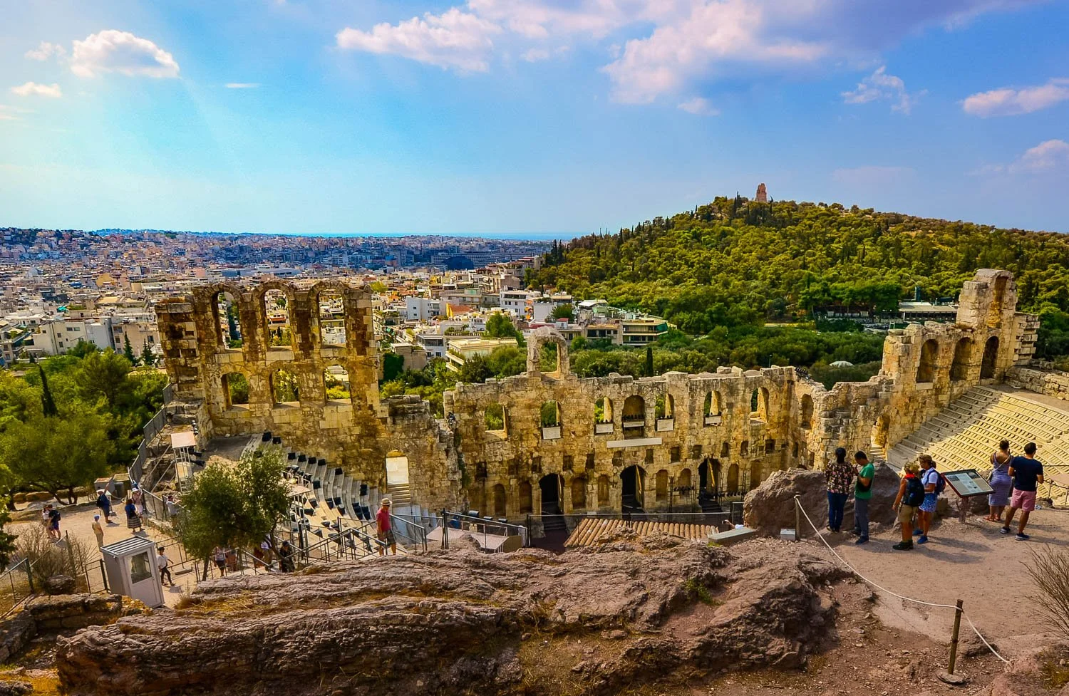 Ancient Greek theater ruins on a hill with a city and a mountain in the background, showing a blue sky with some clouds.
