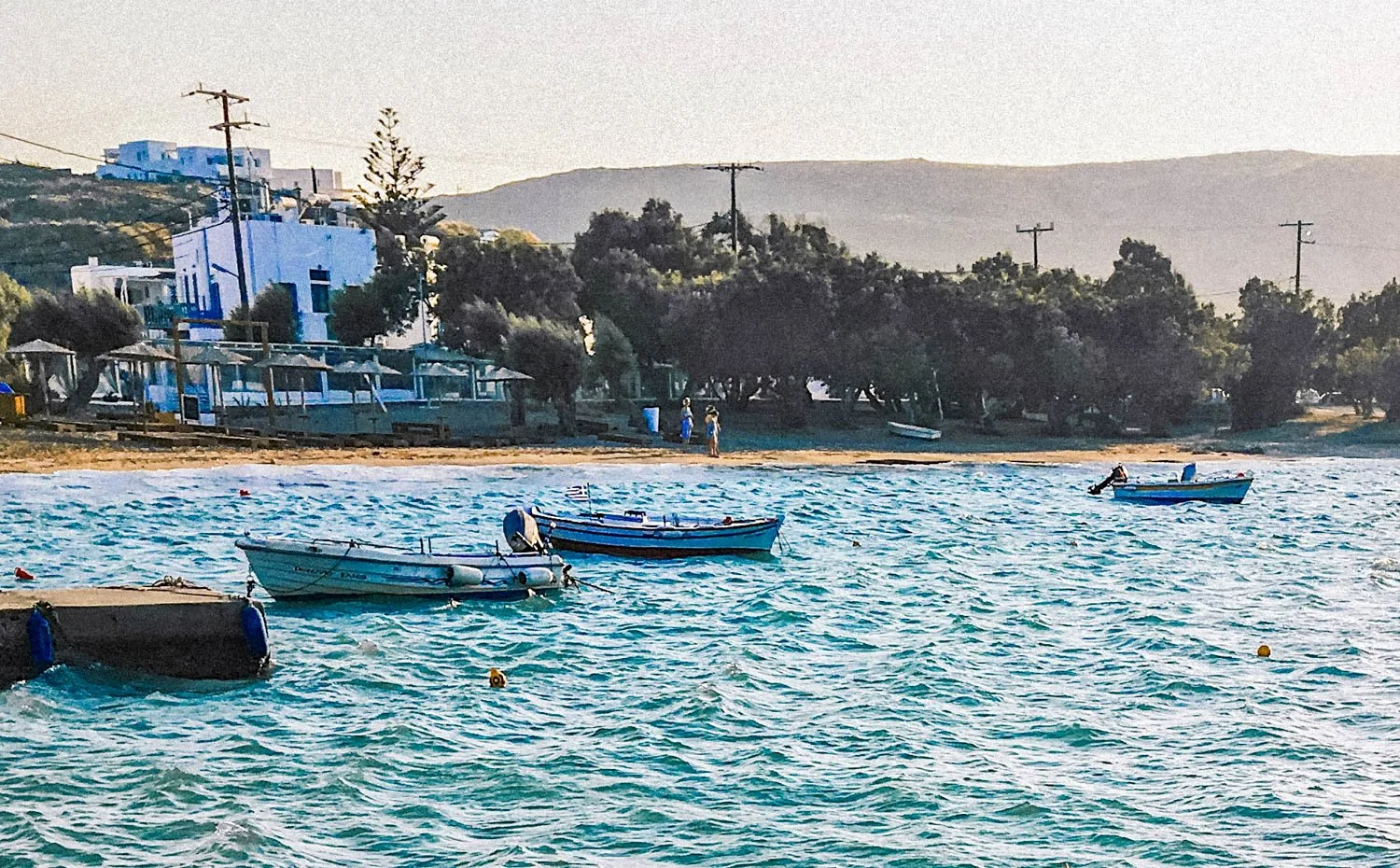 Boats floating on the water near a beach with trees, buildings, and hills in the background.