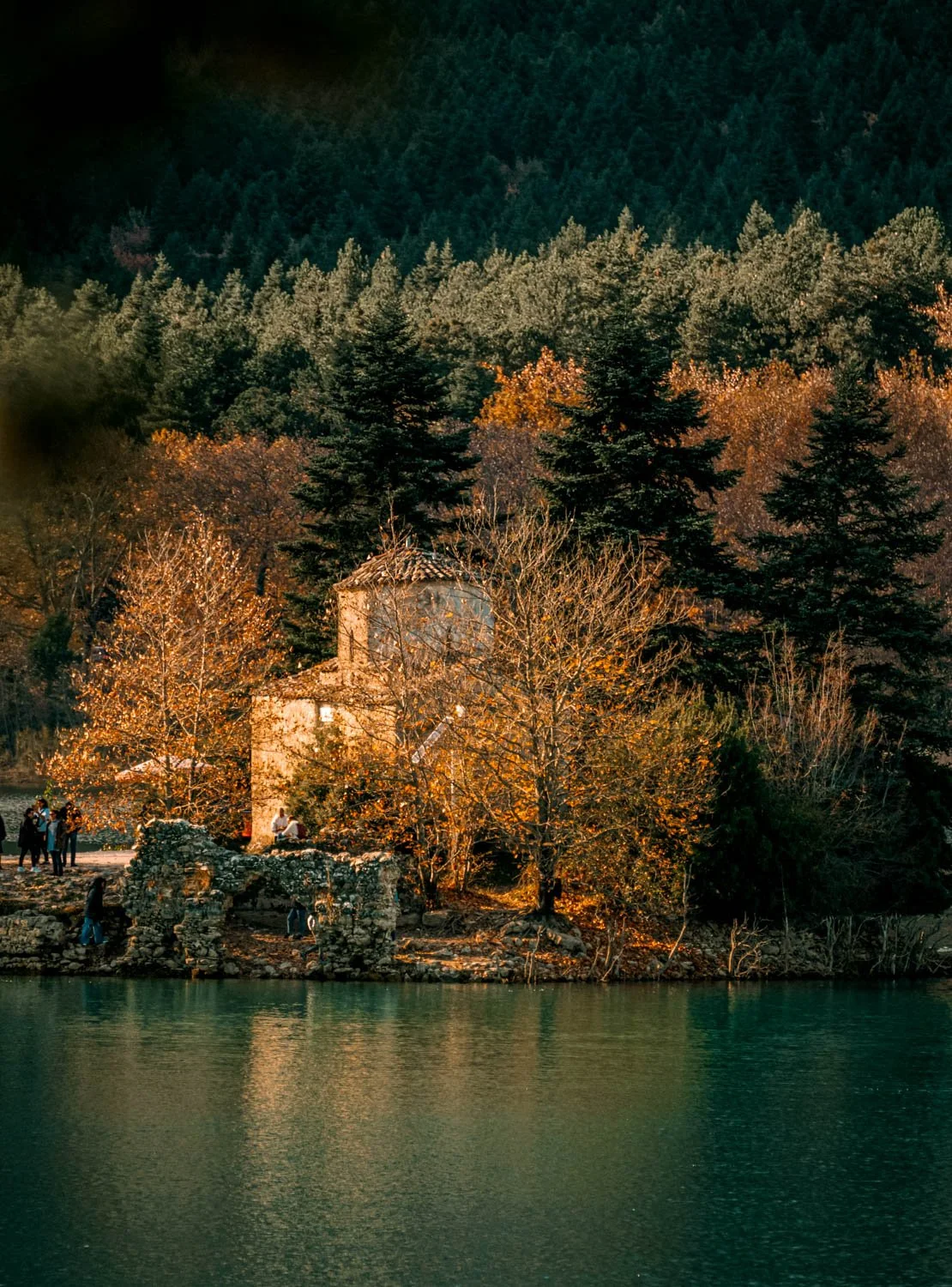 A scenic landscape featuring a small stone building with a tiled roof, surrounded by autumn trees with orange and yellow leaves, located by a calm body of water, with a forested hillside in the background.