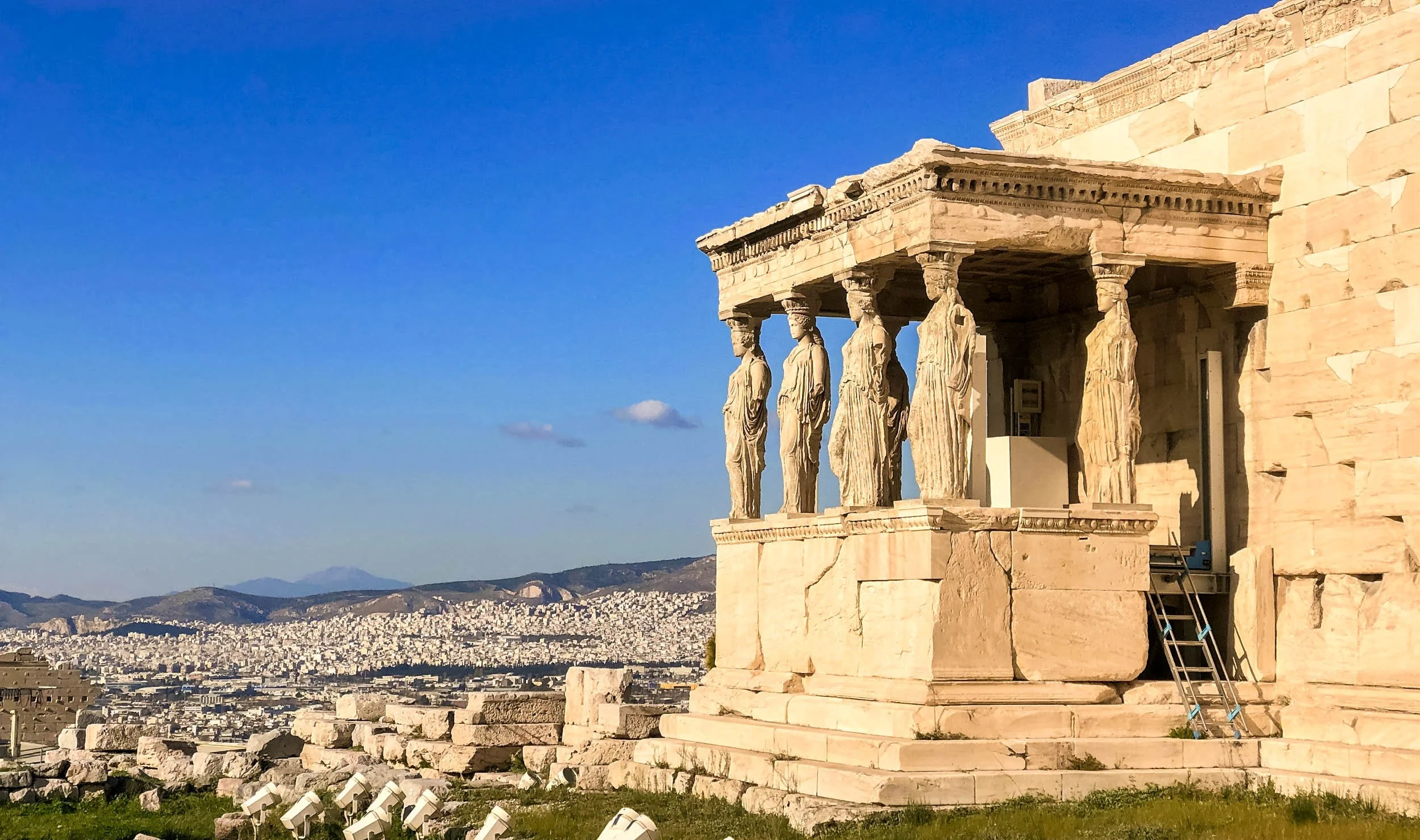 Ancient Greek temple with five caryatid statues supporting the roof, overlooking a city with mountains in the background, under a clear blue sky.