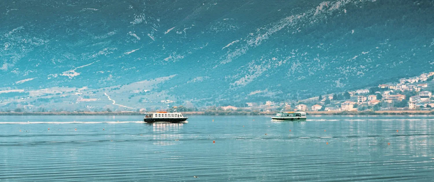 Two boats sailing on a calm lake with a mountainous backdrop and a residential area along the shoreline.