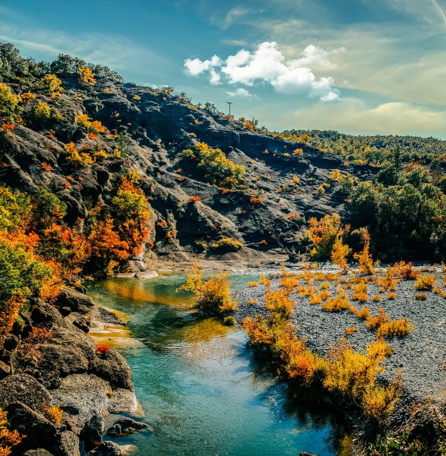 Scenic landscape of a river flowing through a valley with rocky hillsides covered in autumn foliage and trees, under a partly cloudy sky.