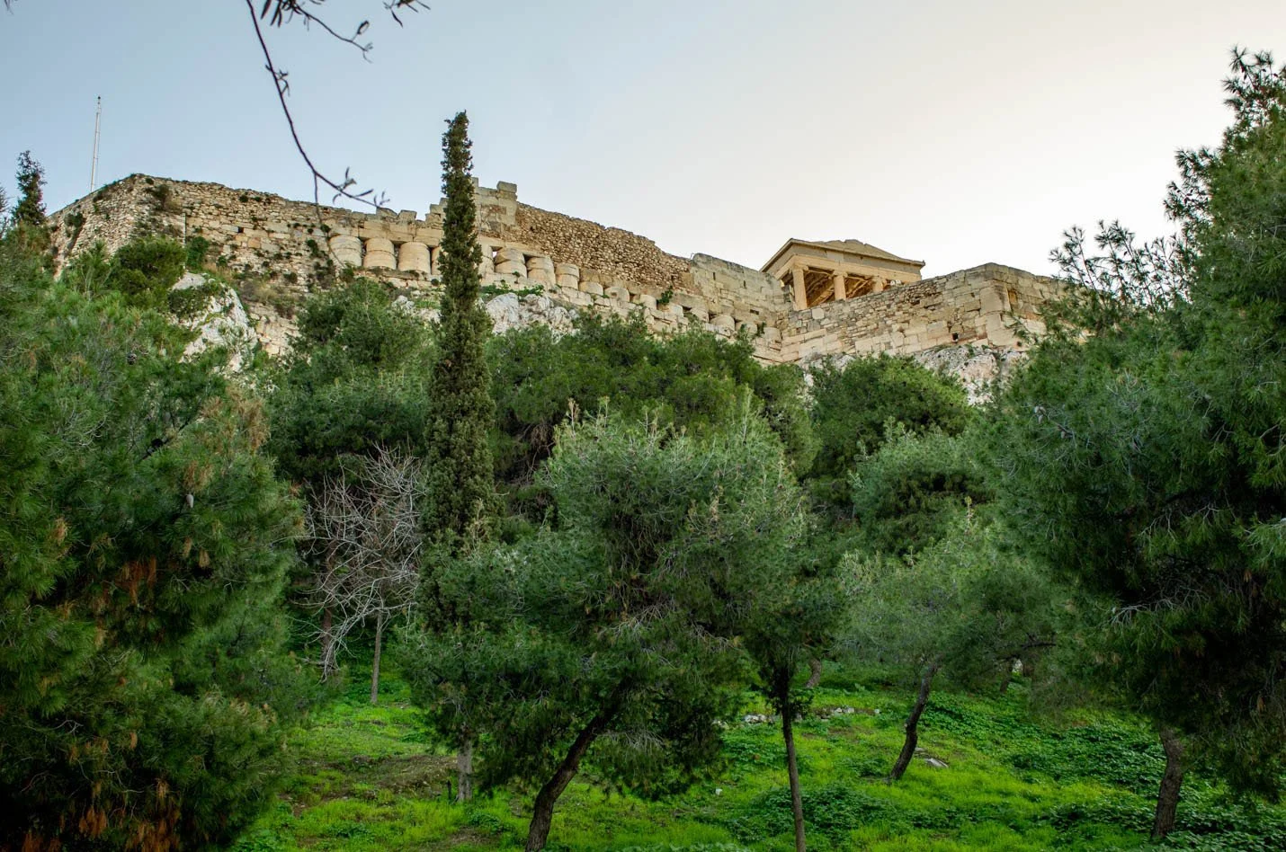 Ancient stone ruins on a hilltop above green trees and bushes under a clear sky.