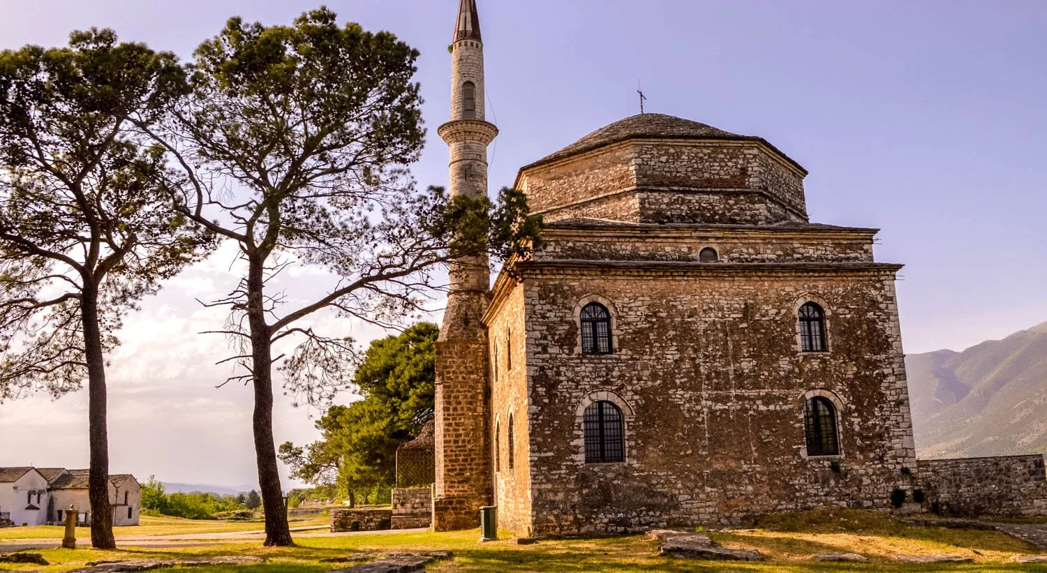 An old stone church with a tall steeple amid surrounding trees and green landscape, mountain range in the background, under a partly cloudy sky.
