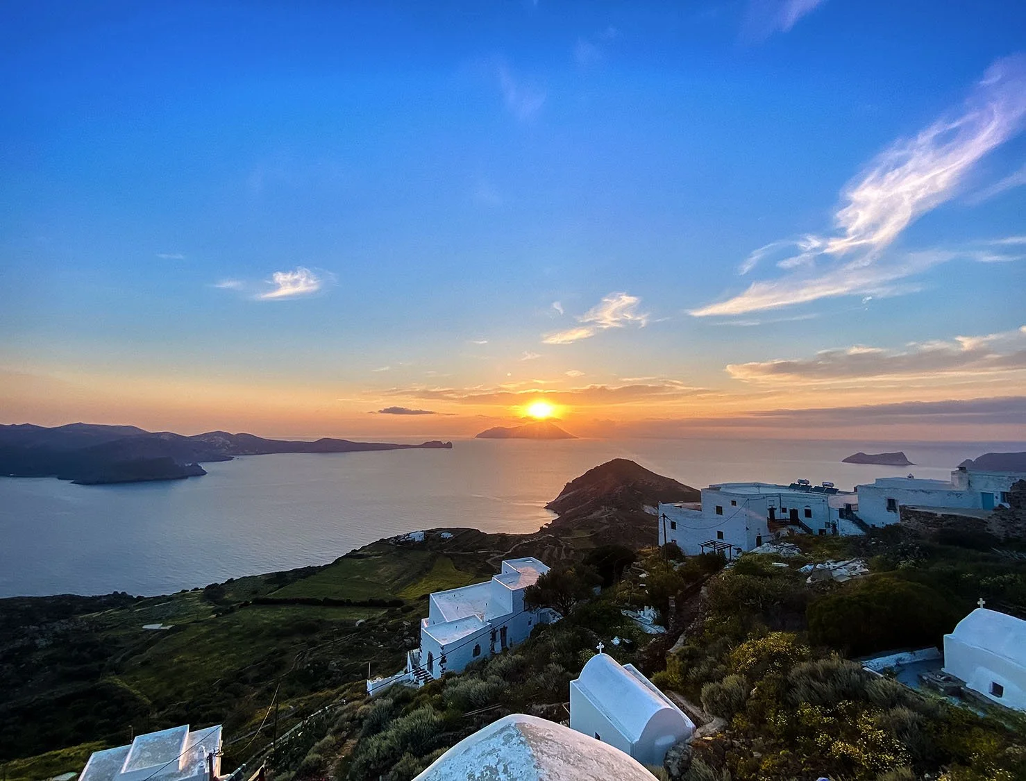 Sunset over a Greek island with white buildings and a large body of water in the background.