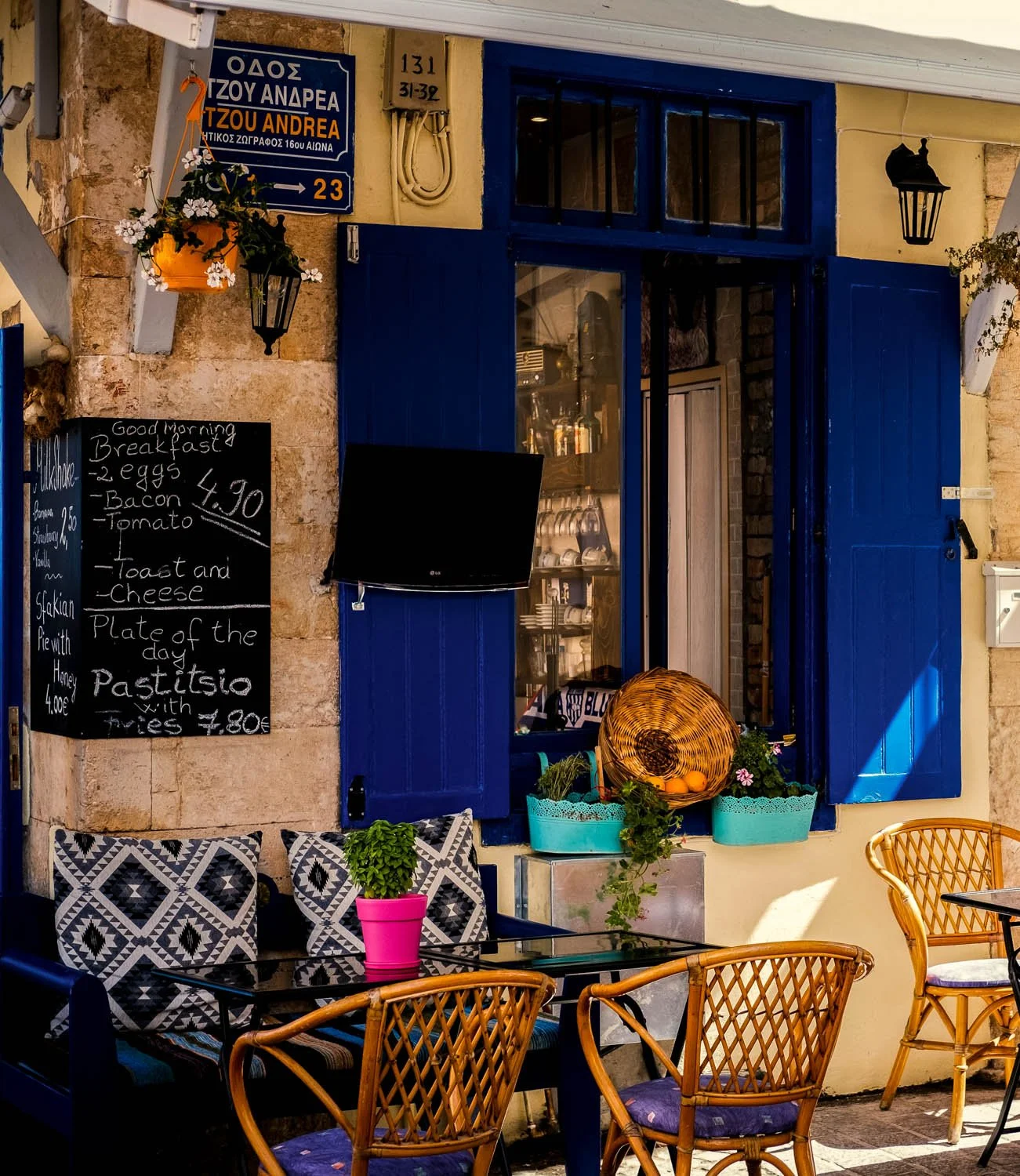 Outdoor seating area of a cafe with patterned cushions, a black table with pink pot plant, wicker chairs, blue window shutters, a blackboard with breakfast menu, and a wicker basket with oranges and flowering plants outside.