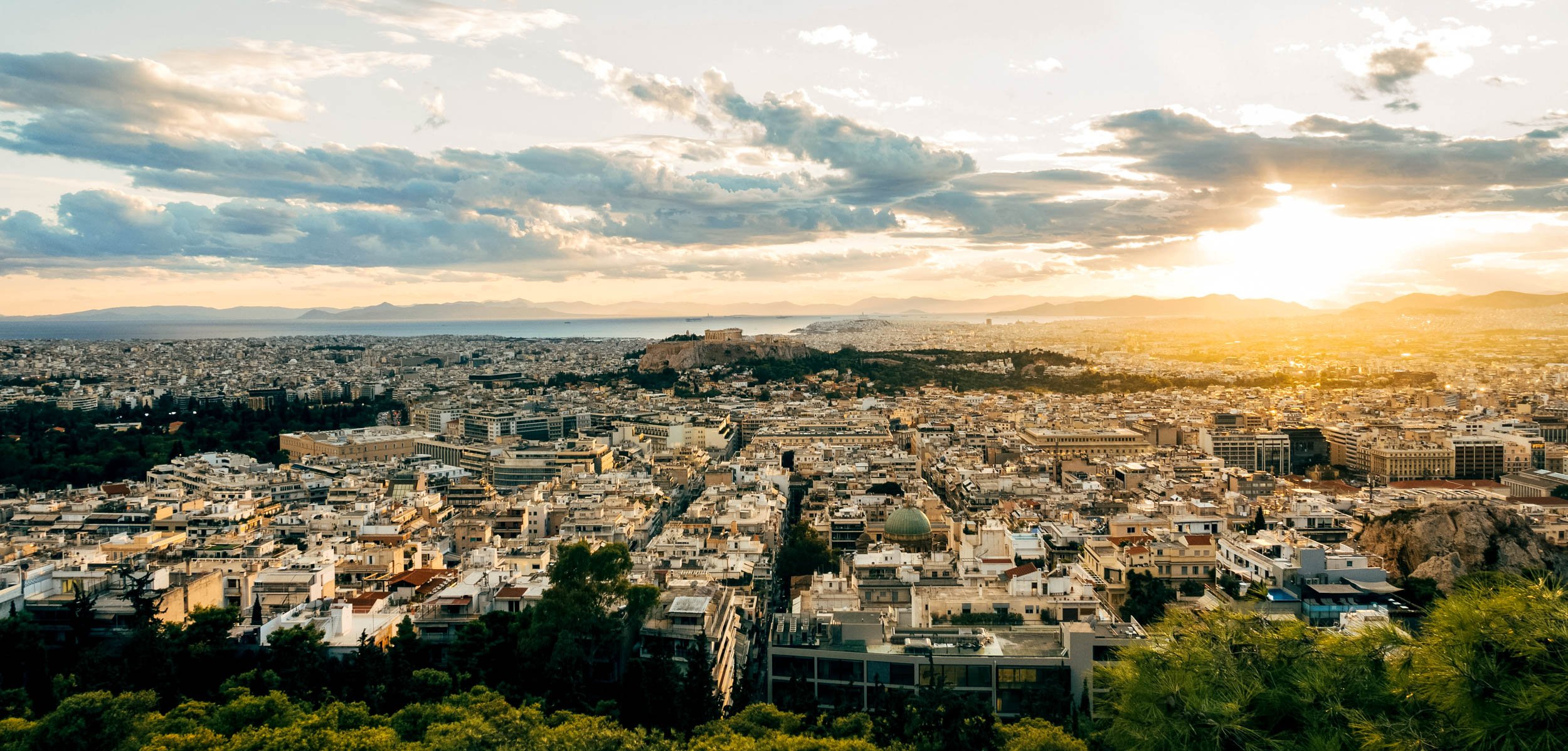 Cityscape of Athens, Greece with ancient Acropolis in the distance during sunset, overlooking white and beige buildings with green trees in the foreground.