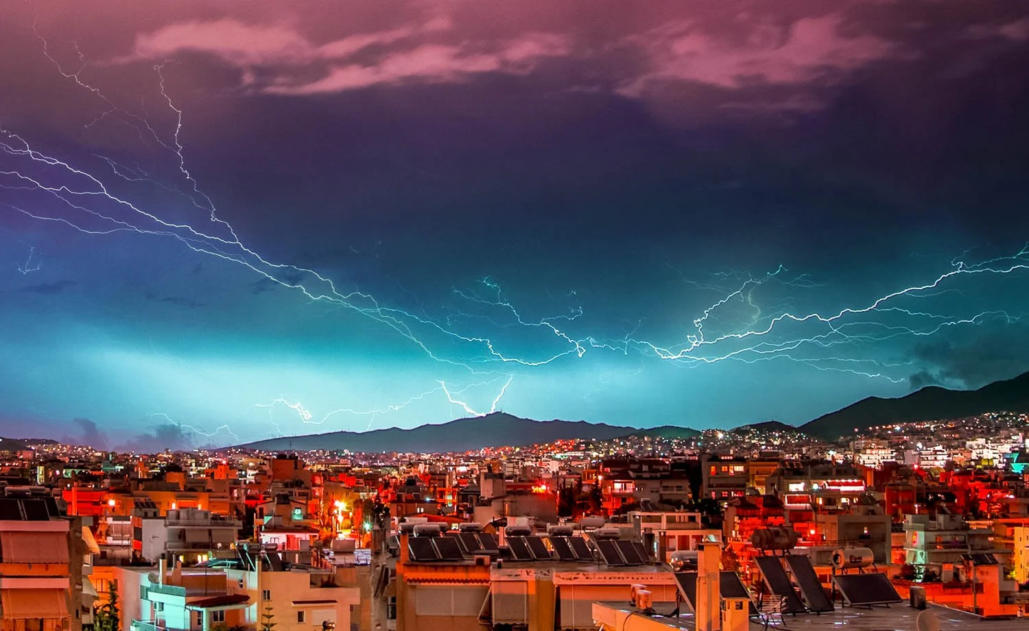 Lightning illuminating a dark stormy sky over a city with mountains in the background at night.