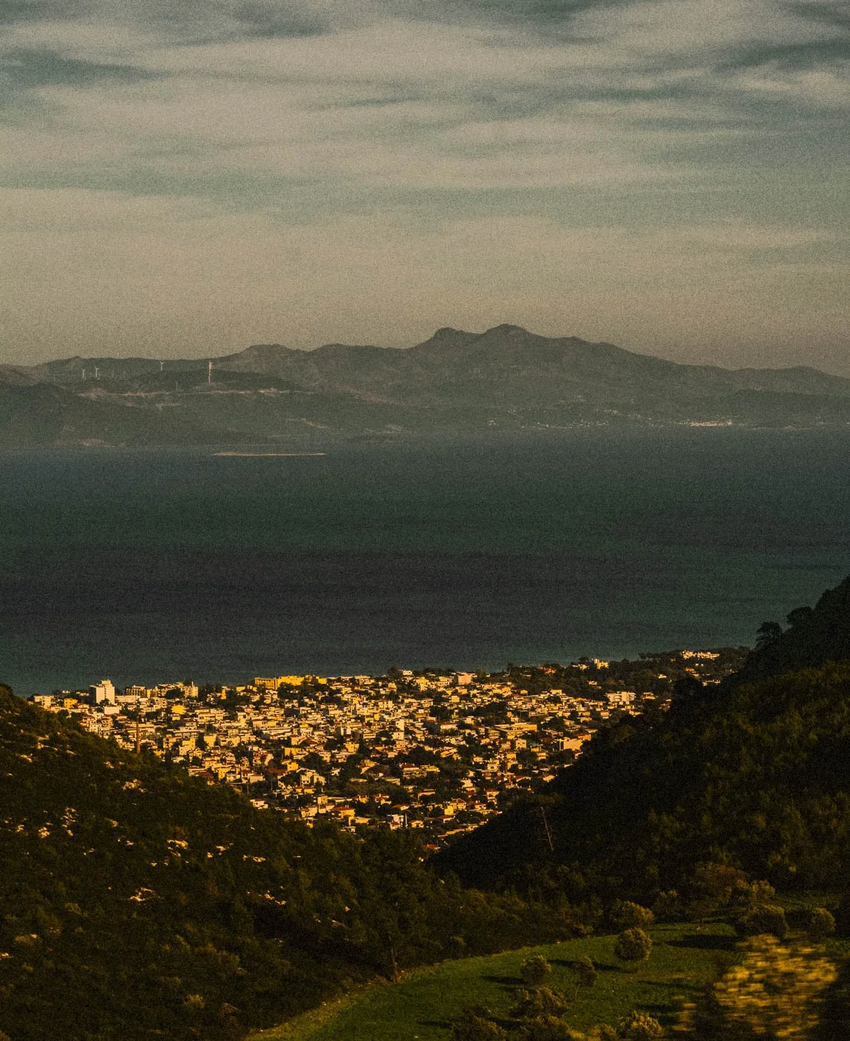 Mountain landscape with a city in the foreground, green hills, and wind turbines on distant peaks.