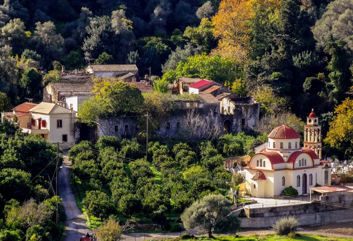A hillside scene with a church featuring white walls and a red roof in the foreground, surrounded by green trees and bushes. More trees and old buildings with tiled roofs are in the background, set against a forested hill.