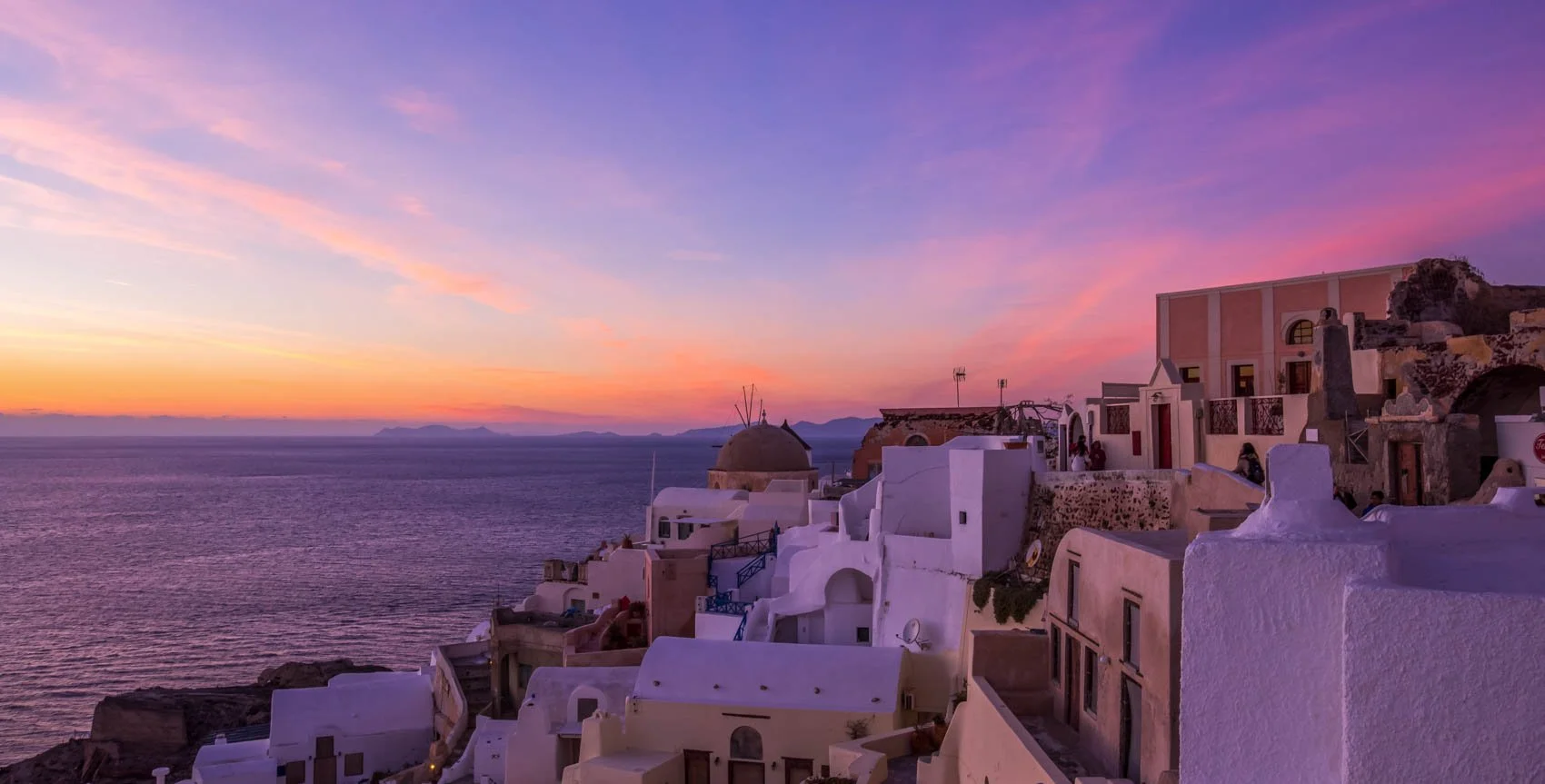 Scenic view of Santorini, Greece at sunset with white buildings and pink, purple, and orange sky over the Aegean Sea.