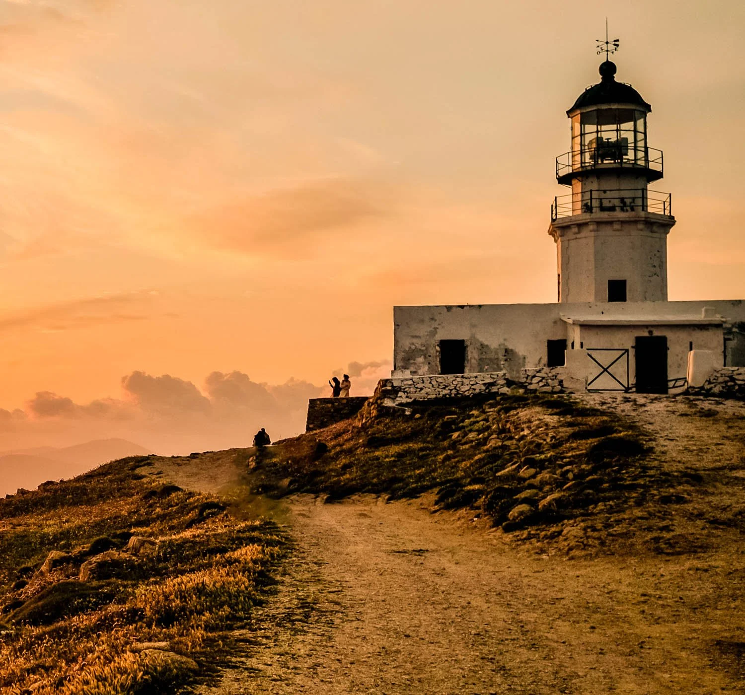 A lighthouse on a hill during sunset with orange and pink sky, two people taking a photo near the lighthouse, and a person sitting on the ground along a dirt path.