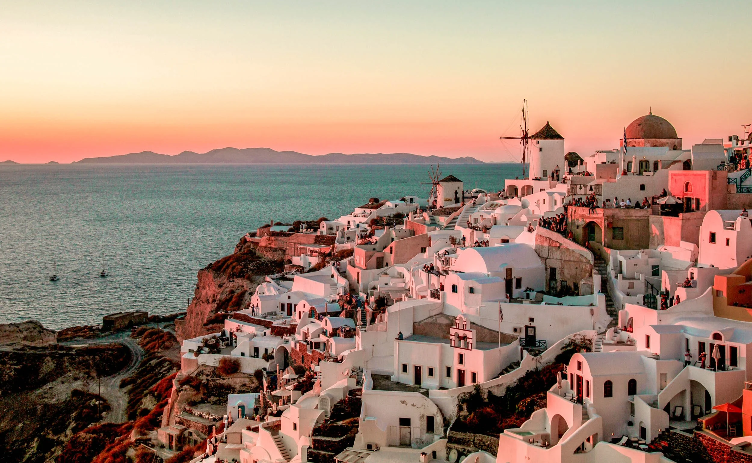 Sunset over white buildings and windmills on the cliffs of a Greek island, with the Aegean Sea in the background.