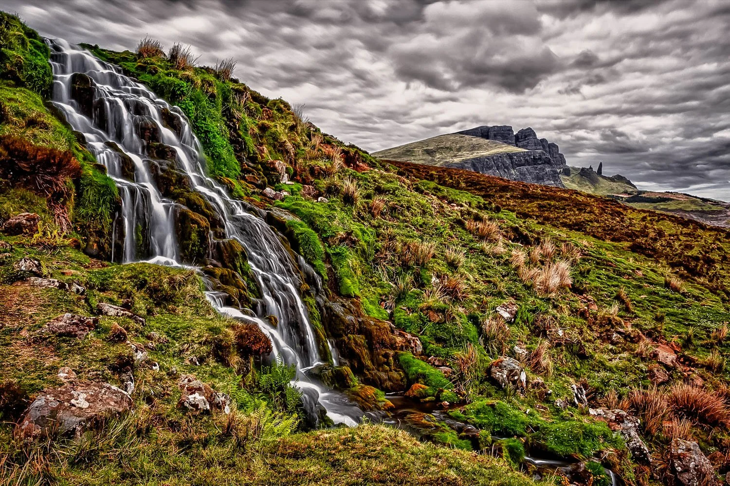 A cascading waterfall flowing down a moss-covered hillside in a rugged, green landscape with mountains in the background under a cloudy sky.