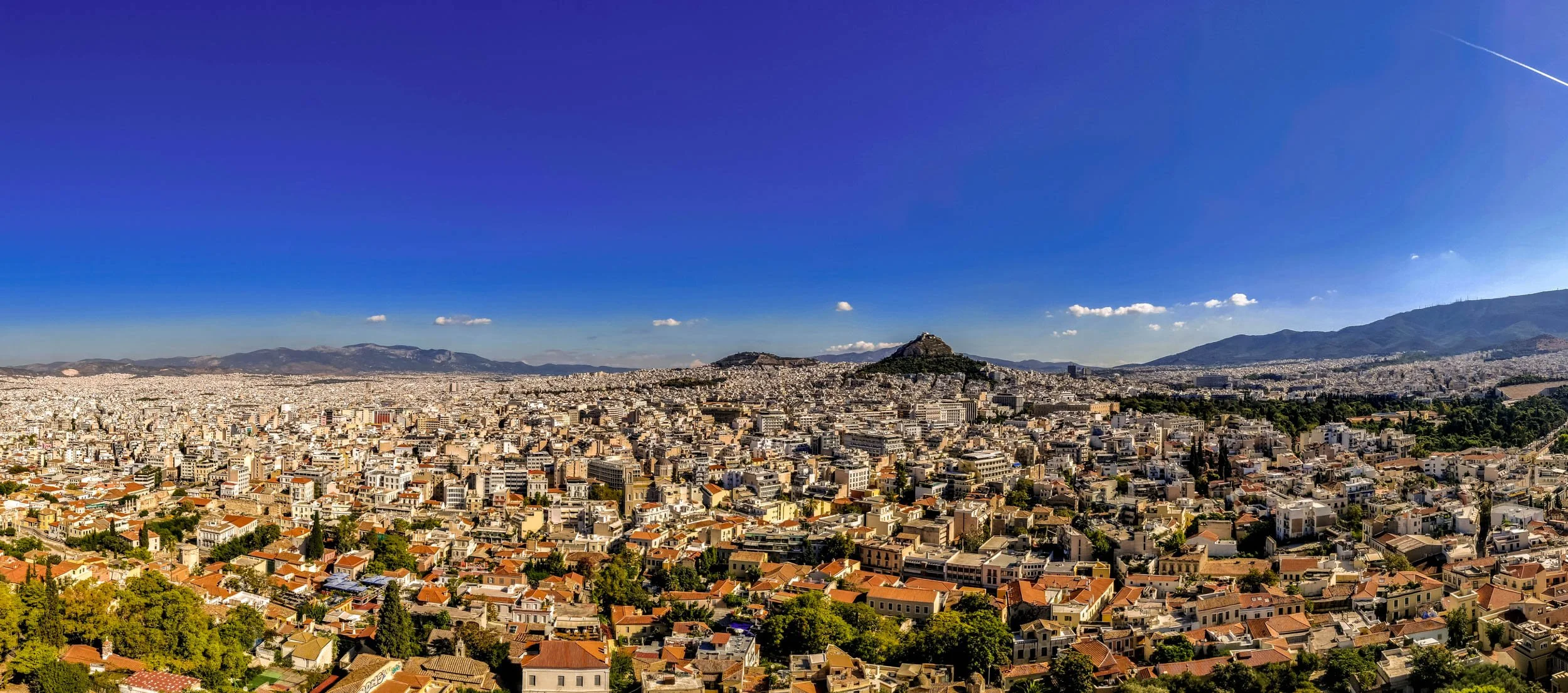 A panoramic view of Athens, Greece, with numerous buildings and houses in the foreground, the Acropolis hill with the Parthenon on top in the middle, surrounded by mountainous terrain under a clear blue sky with a few clouds.