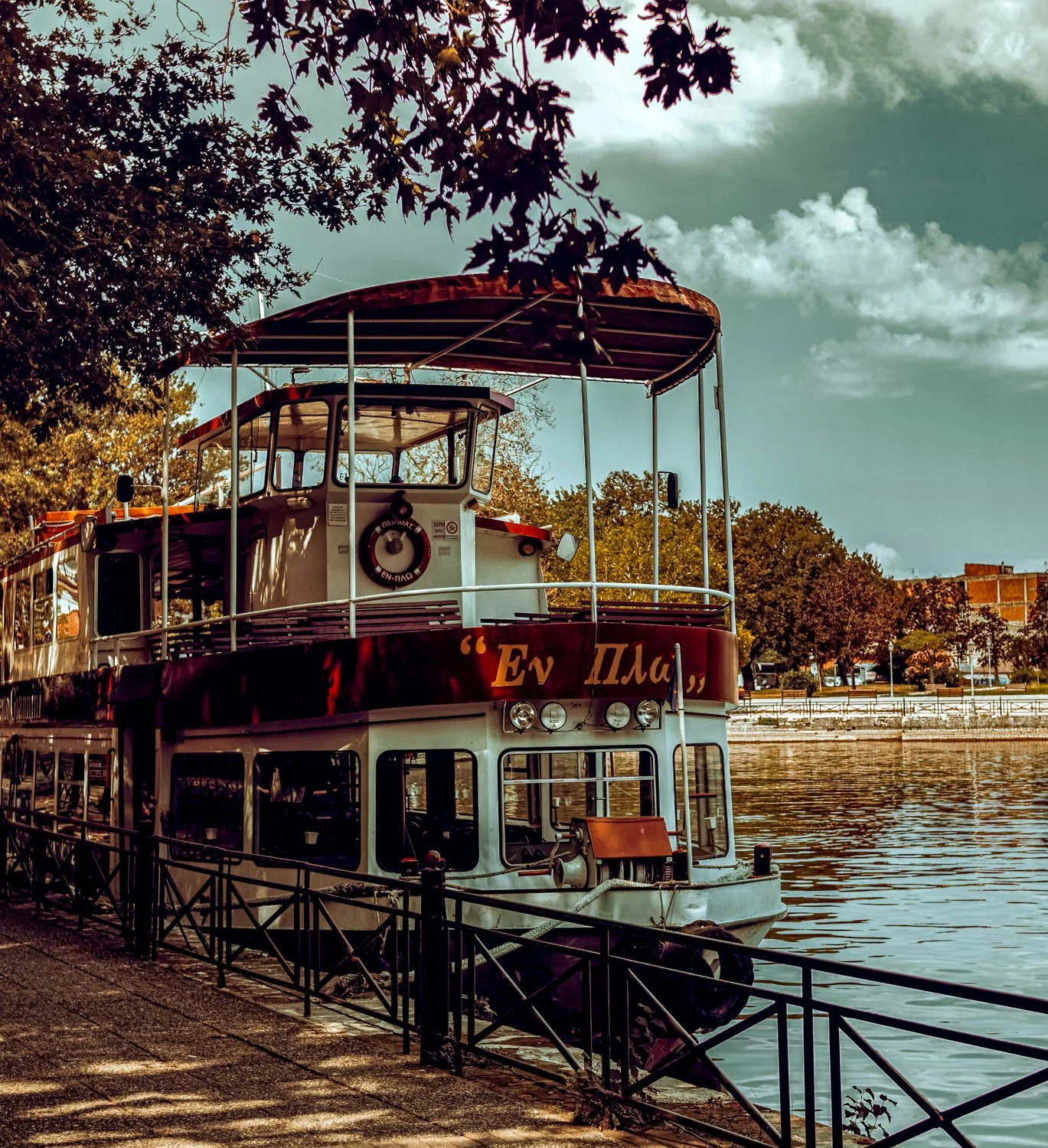A boat docked on a river, partially shaded by tree branches, with a waterway and trees in the background.