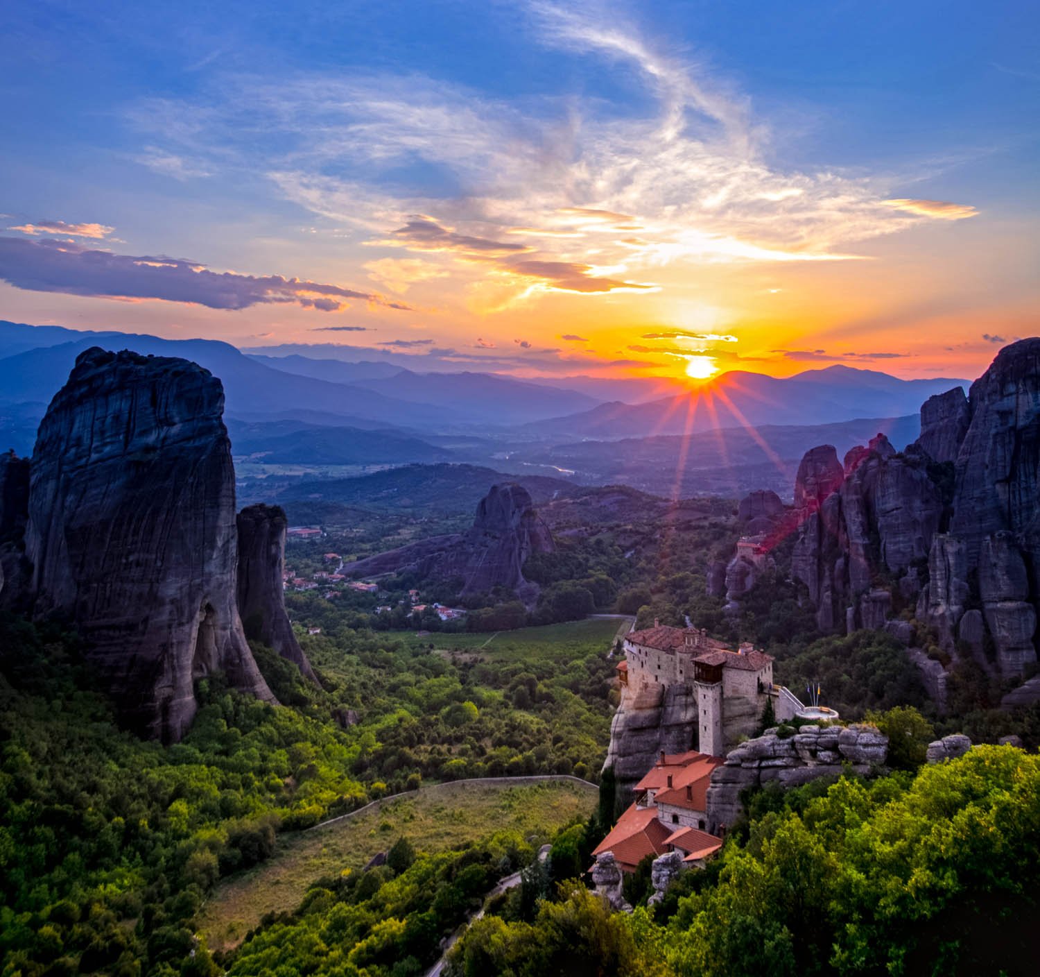 Sunset over rocky mountains and a monastery perched on a cliff in Meteora, Greece, with a valley and distant mountains in the background.