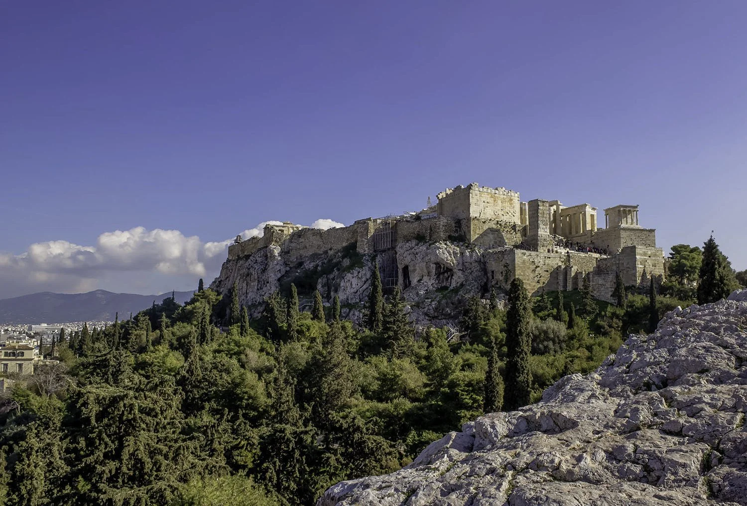 Ancient acropolis atop a rocky hill with tall trees and modern buildings in the background, under a blue sky with some clouds.