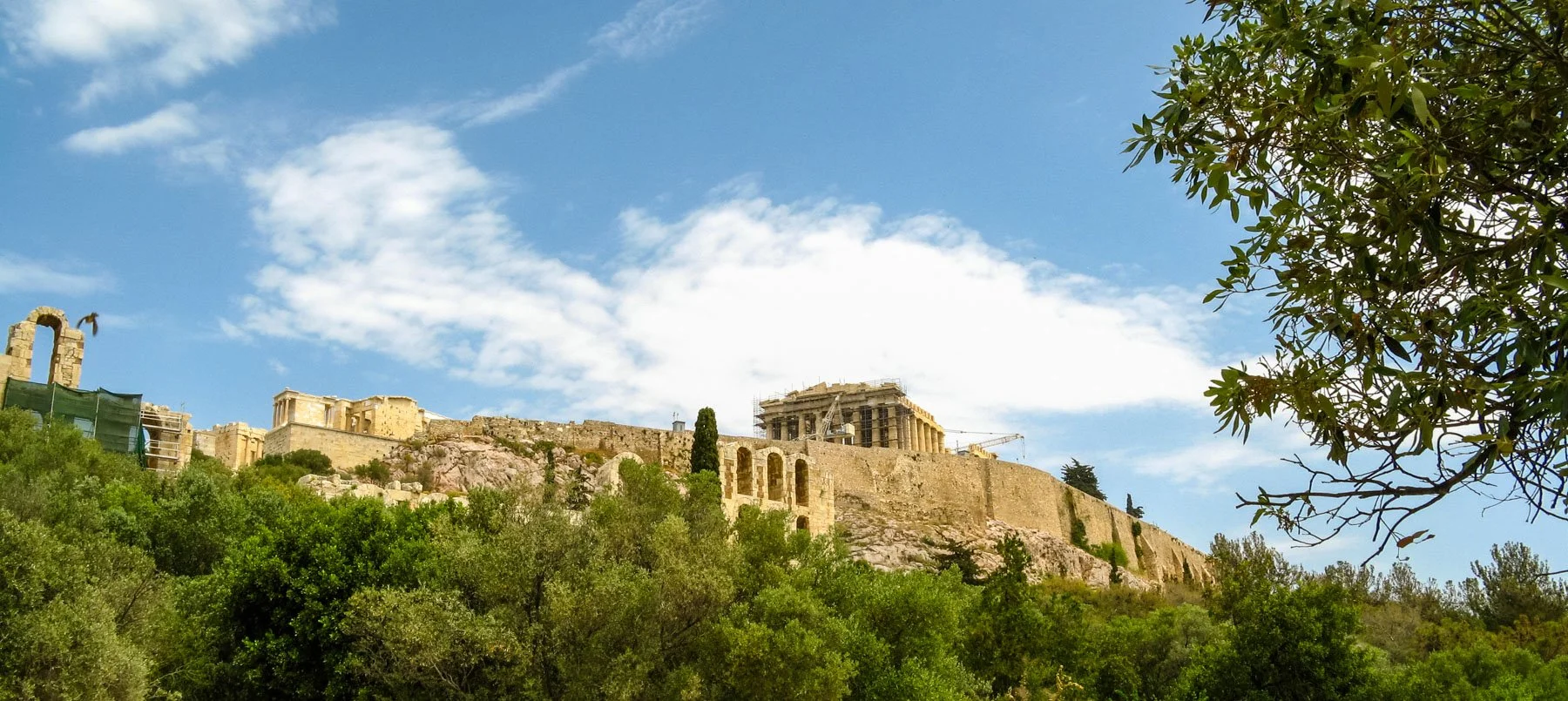 Ancient ruins of the Acropolis on a hilltop with a partially cloudy sky in the background and trees in the foreground.