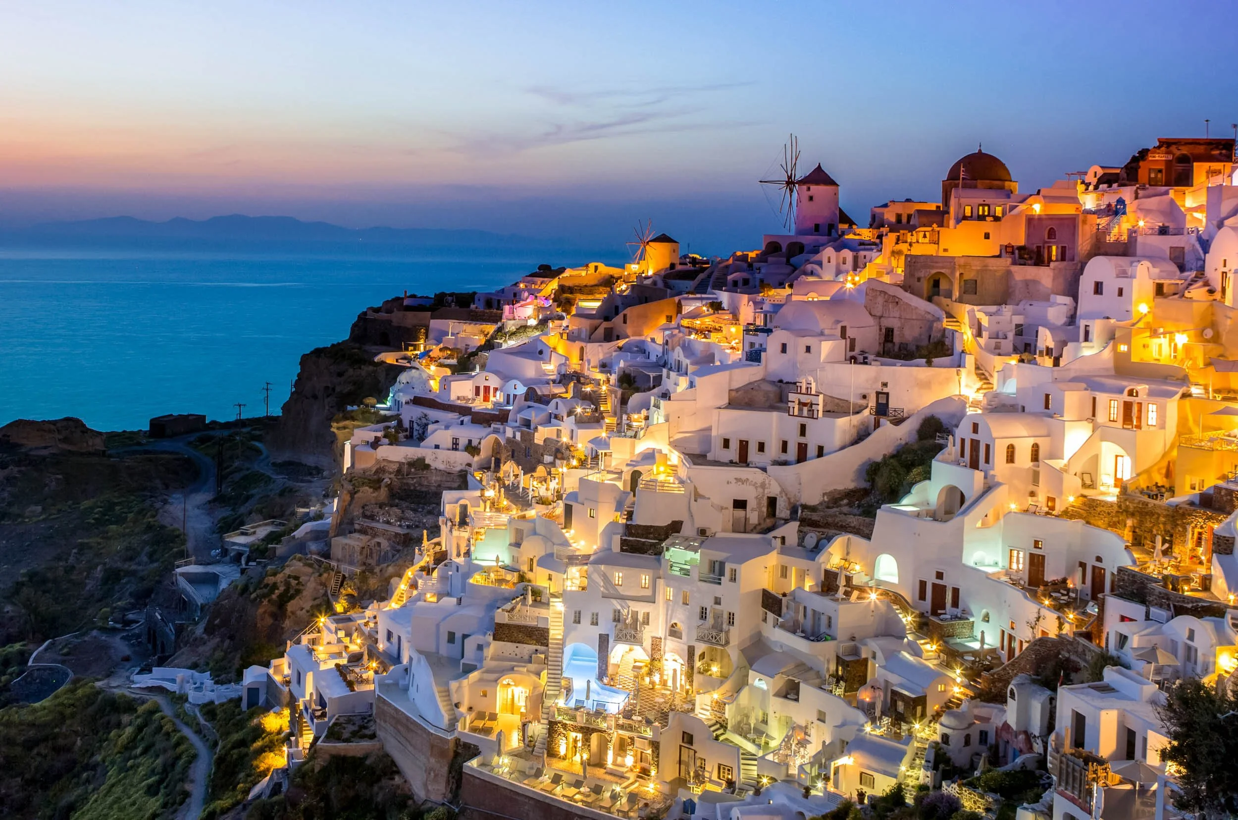 Photograph of Santorini, Greece at sunset showing white buildings with blue domes on a hillside overlooking the sea.