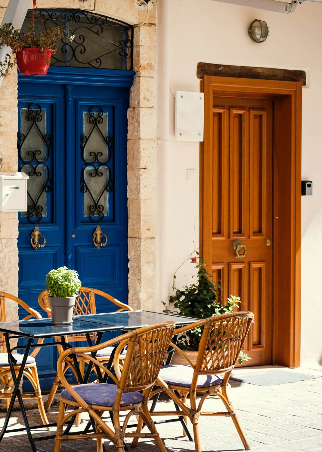 Outdoor patio with a glass table and rattan chairs, green potted plant on the table, blue door with black wrought iron accents, wooden door, and potted plant with red flowers.