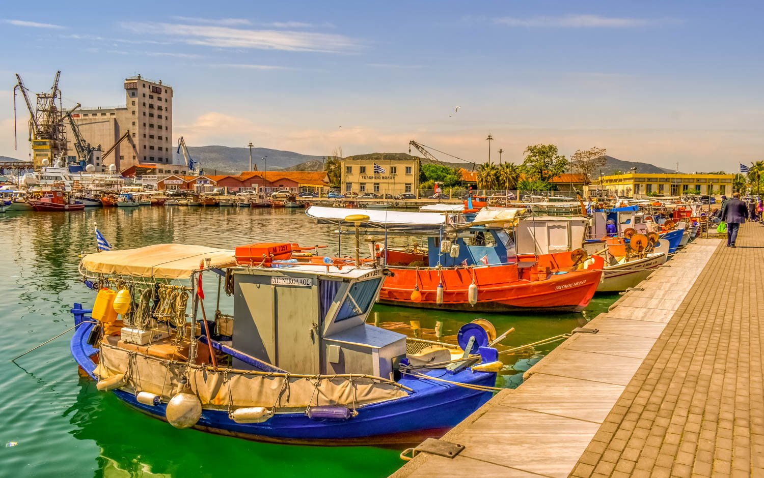 Boats docked at a harbor with colorful buildings and hills in the background on a sunny day.
