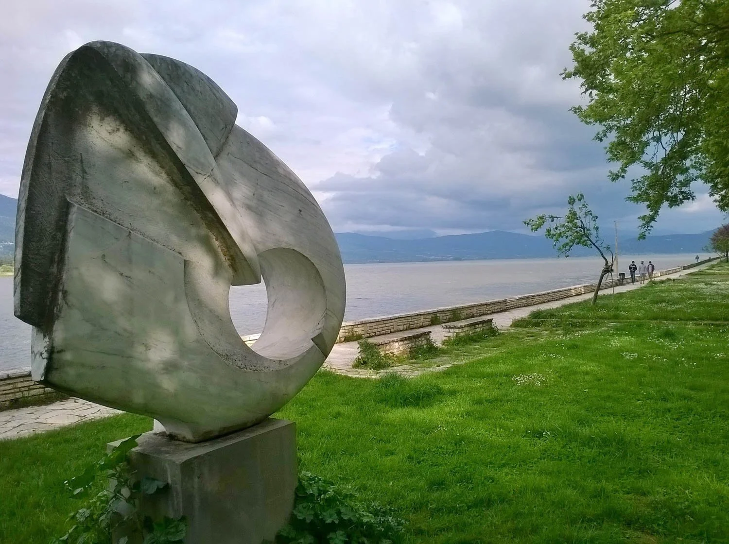 Large abstract metal sculpture by a lake with a tree and people walking on a pathway in the background, mountains in the distance, and cloudy sky.