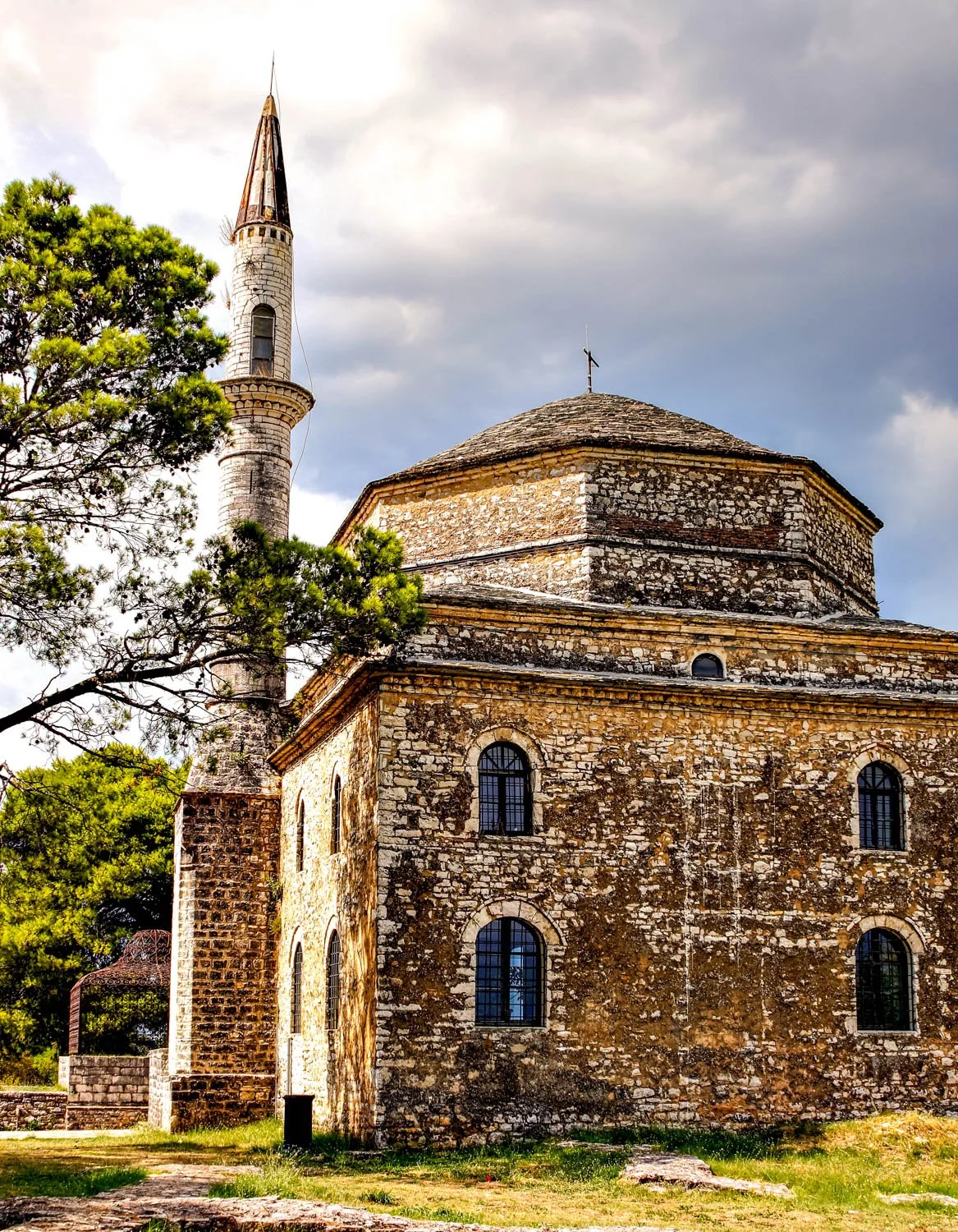 An old stone church with arched windows and a tall, pointed minaret, surrounded by green trees under a cloudy sky.