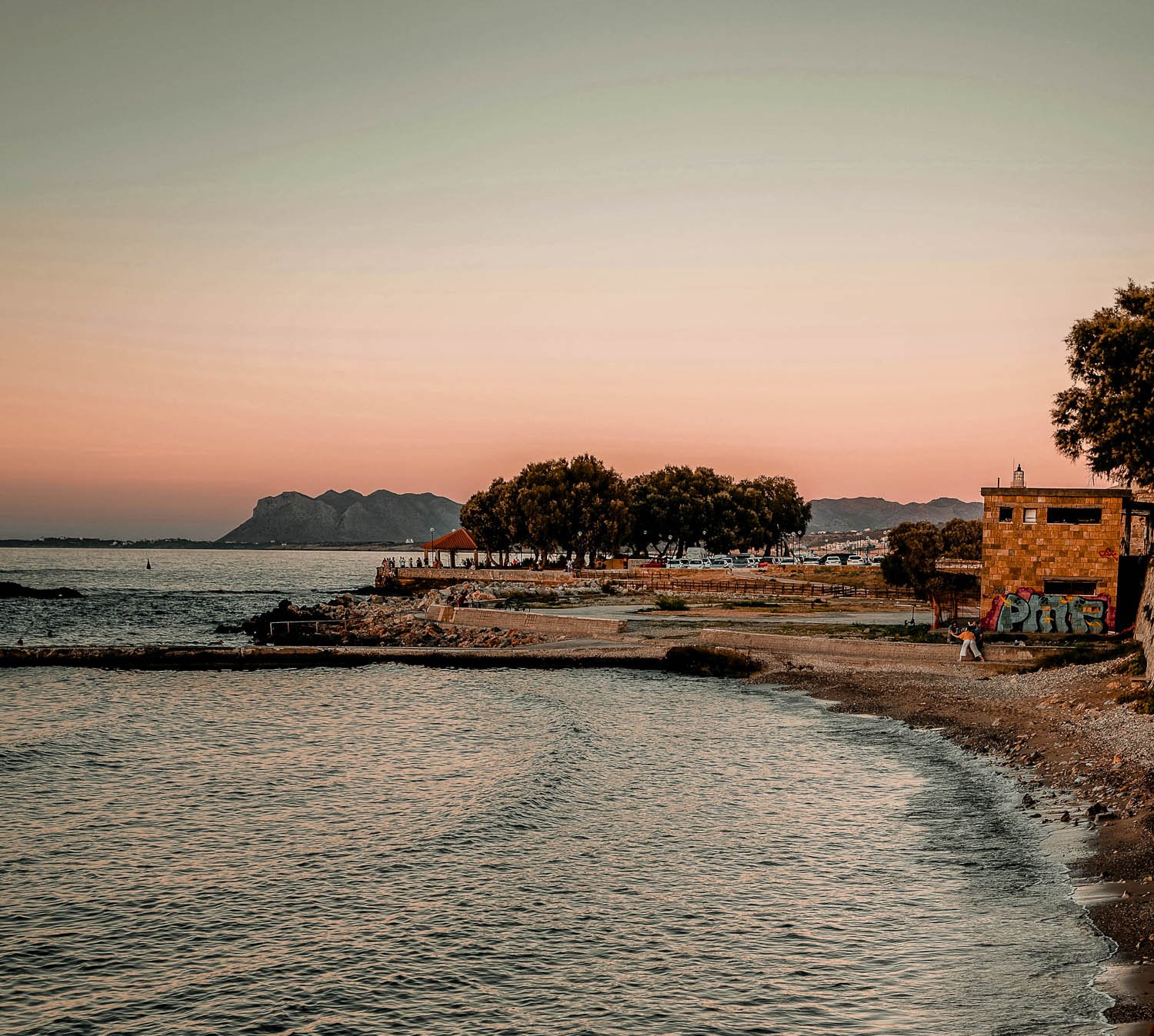 A peaceful beach scene at sunset with calm water, a rocky shoreline, trees, a graffiti-covered building, a mountain in the distance, and a pathway with a person walking.
