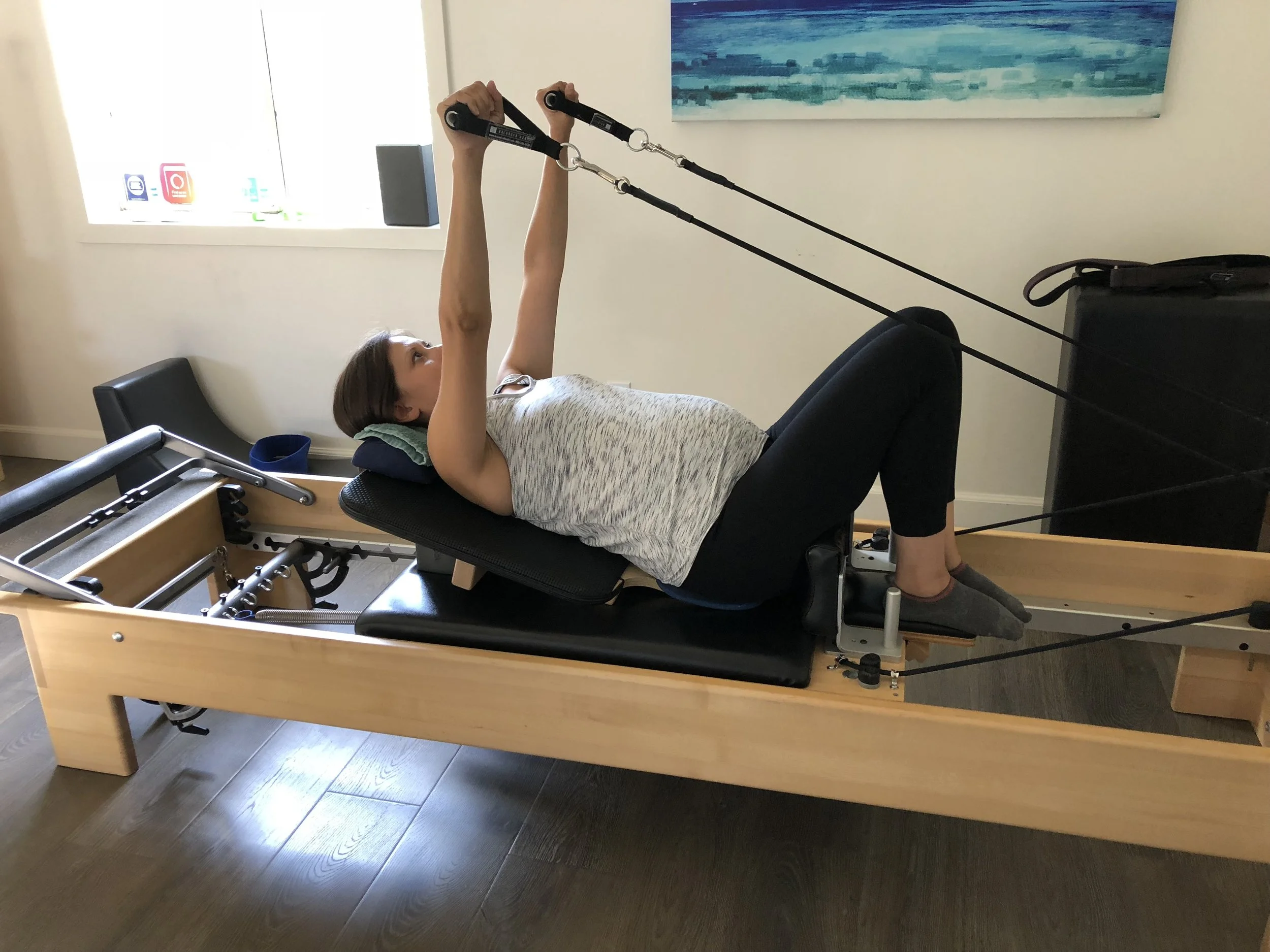Woman lying on a Pilates reformer machine, holding resistance bands above her head, engaging in exercise in a room with a window and artwork on the wall.
