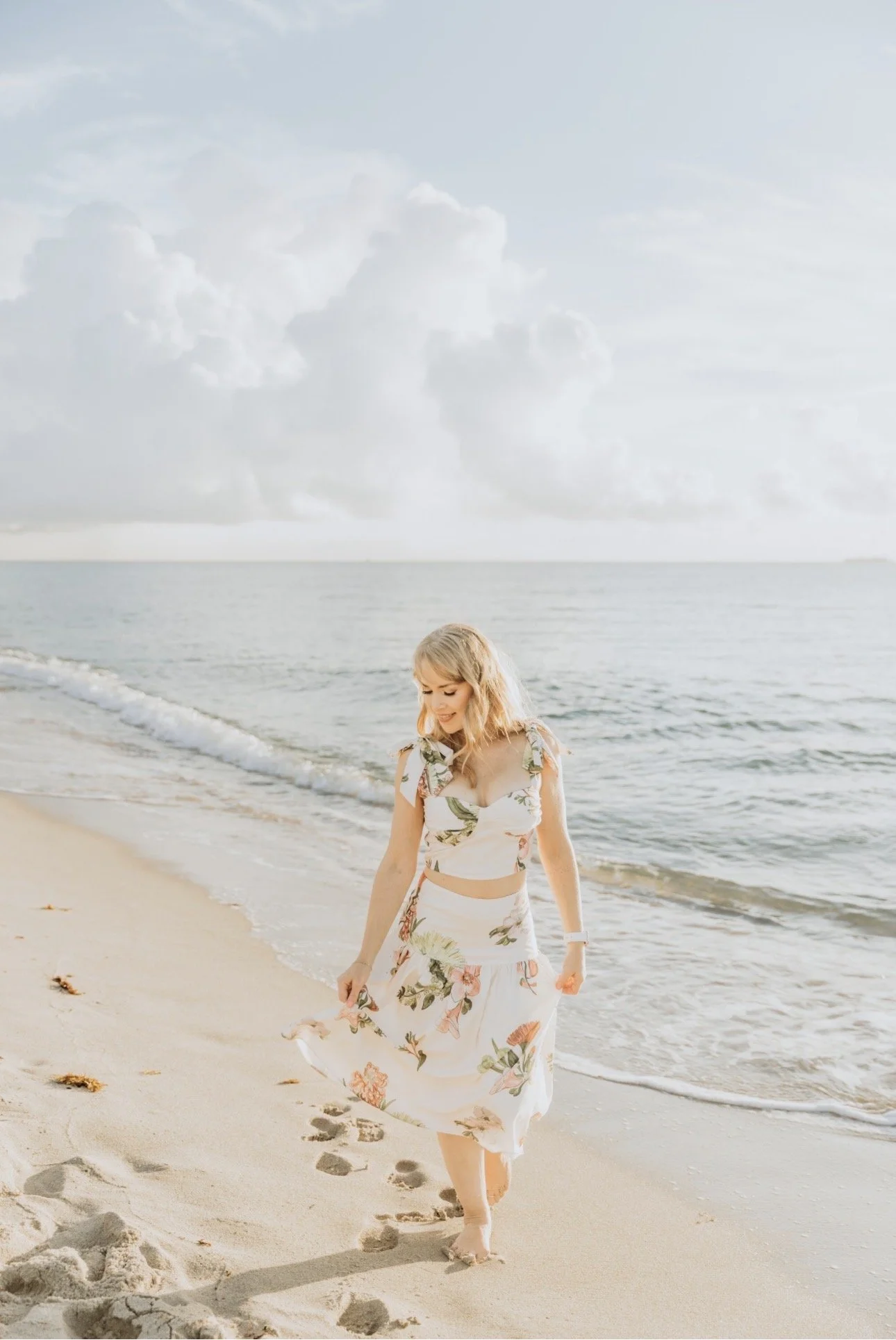 Founder of Ozean Pilates + Wellness is  woman with blonde hair walking along Miami Beach in the morning sunshine, wearing a floral dress, smiling, with footprints visible in the sand.