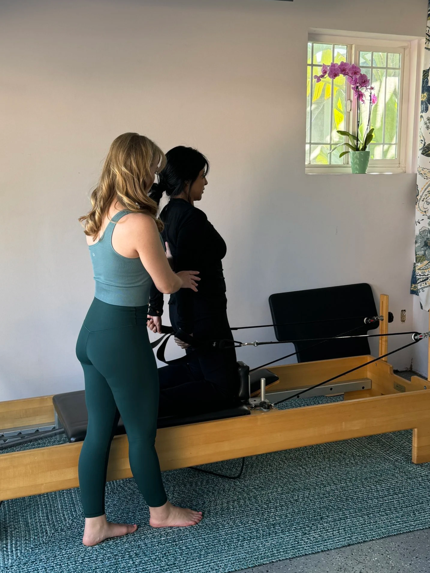 A female client in her 30's on the Pilates Reformer being cued in the Kneeling Series Chest Expansion exercise in a private Pilates Pre-natal + Postpartum session.