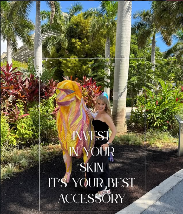 A woman standing outdoors with lush tropical plants and trees around her, wearing a black strapless dress, posing next to a colorful, artistic horse sculpture with pink, yellow, and purple streaks, with the quote 'Invest in your skin. It's your best accessory' overlayed on the image.