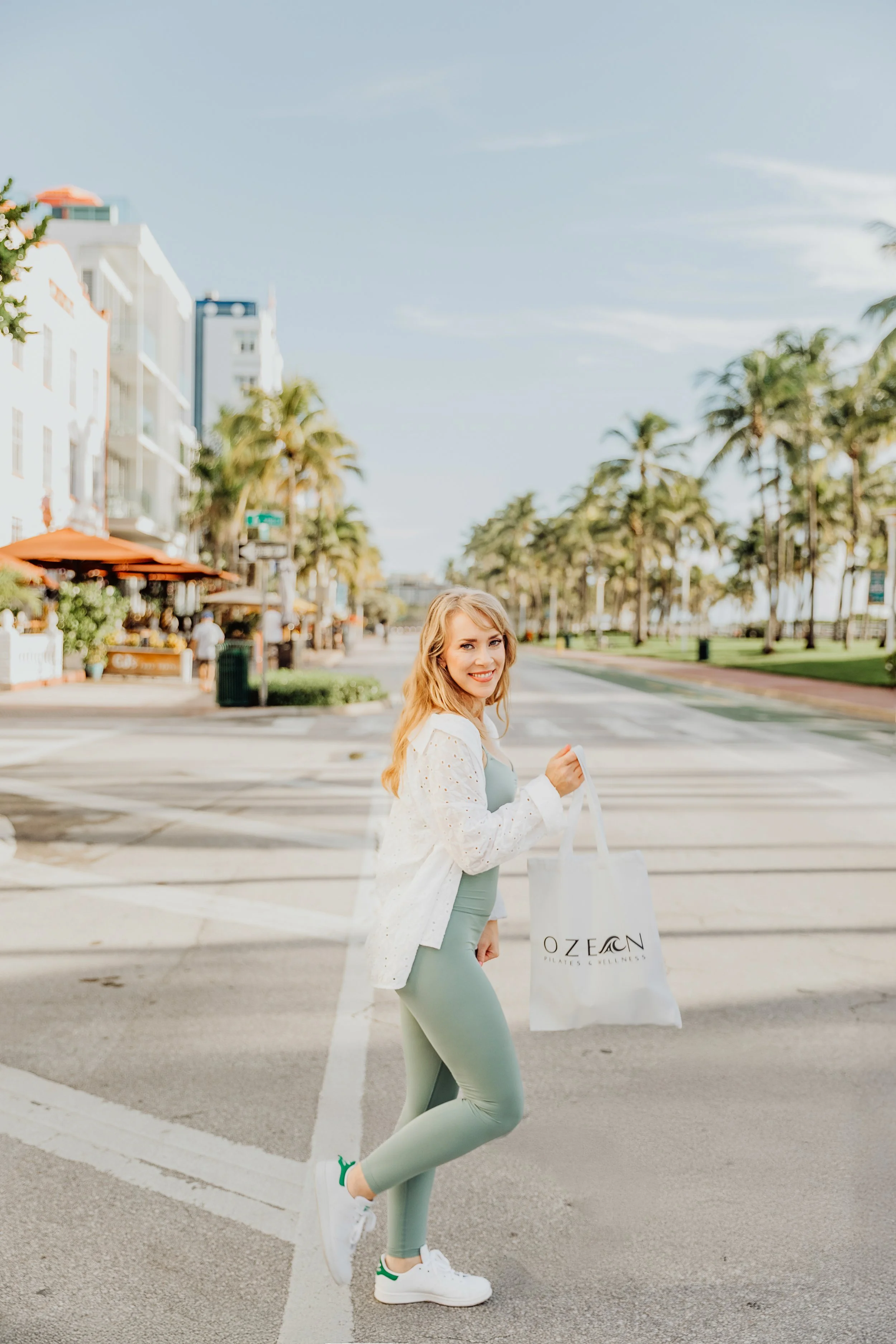 Founder of Ozean, smiling walking on Ocean Drive in Miami Beach  holding an Ozen Pilates & Wellness tote, with palm trees lining the street and outdoor cafes on the sidewalk.