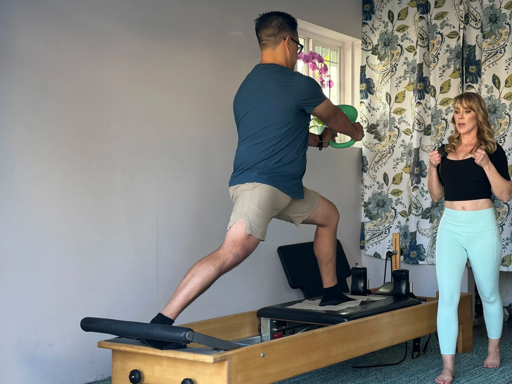 A male client in his 40's is lifting a green kettlebell while performing a standing lunge exercise on the Pilates Reformer in a private Athletic Performance session with Pilates teacher and founder of Ozean cueing and observing him
