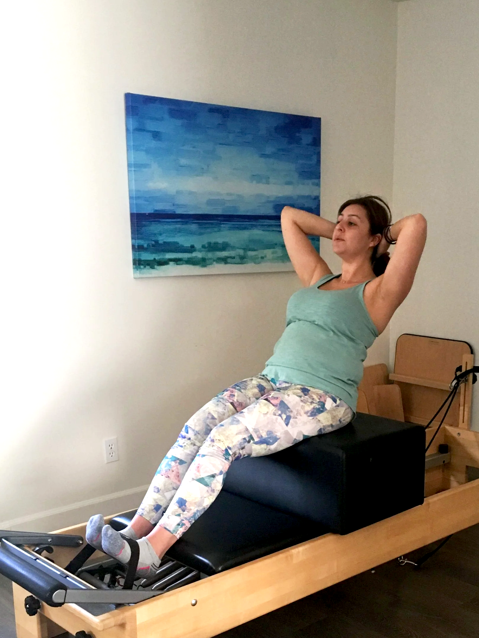A female client in her 30's in a private Pilates Longevity session on Pilates Reformer with her hands behind her head performing Classical Short Box Abs Series with a painting of a blue seascape on the wall.