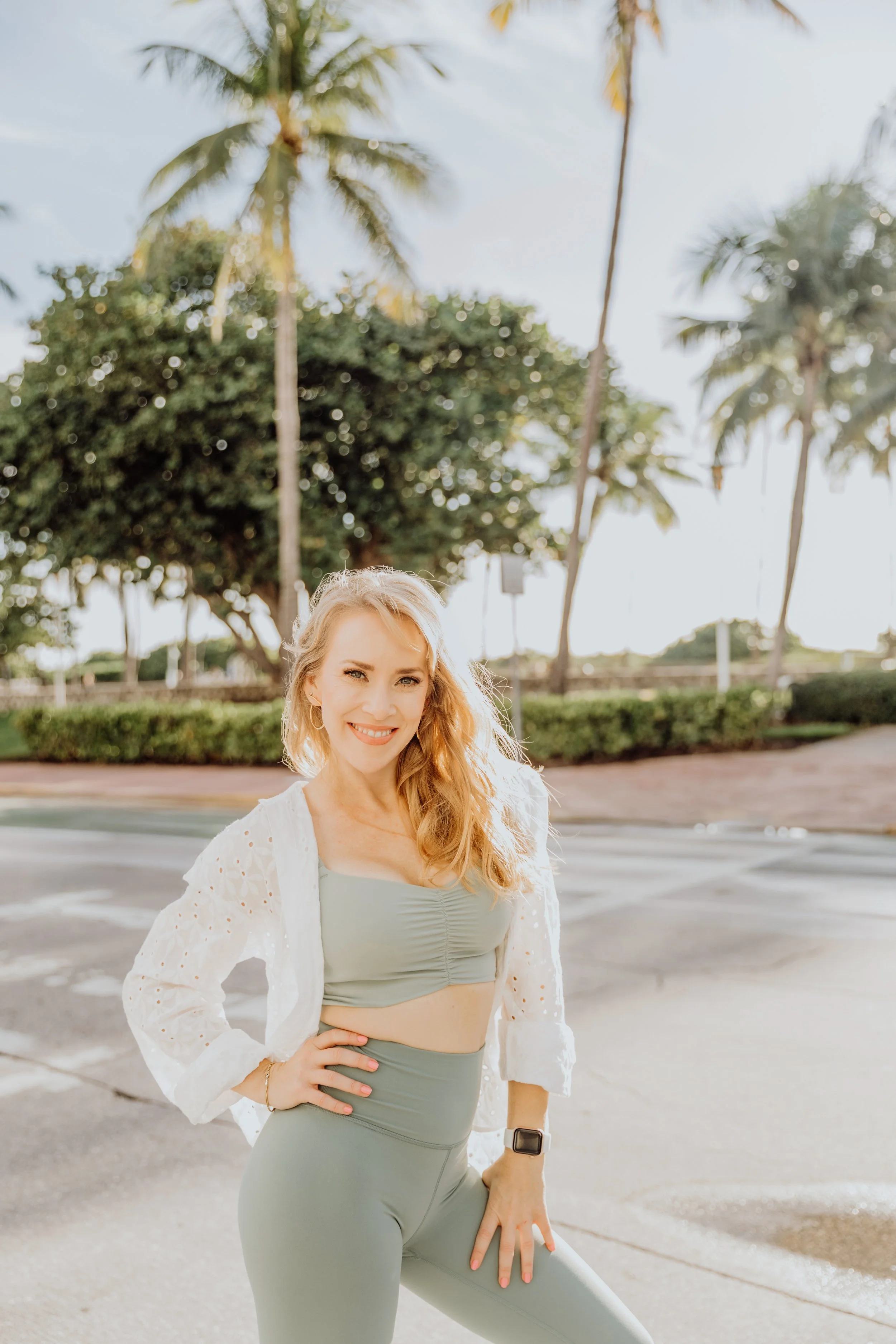 A smiling Pilates Teacher woman with long, wavy blonde hair, wearing a light sage green Athleisure wear, a white eyelet cardigan, and a smartwatch, standing outdoors on a sunny day in Miami Beach with palm trees and a paved area in the background.