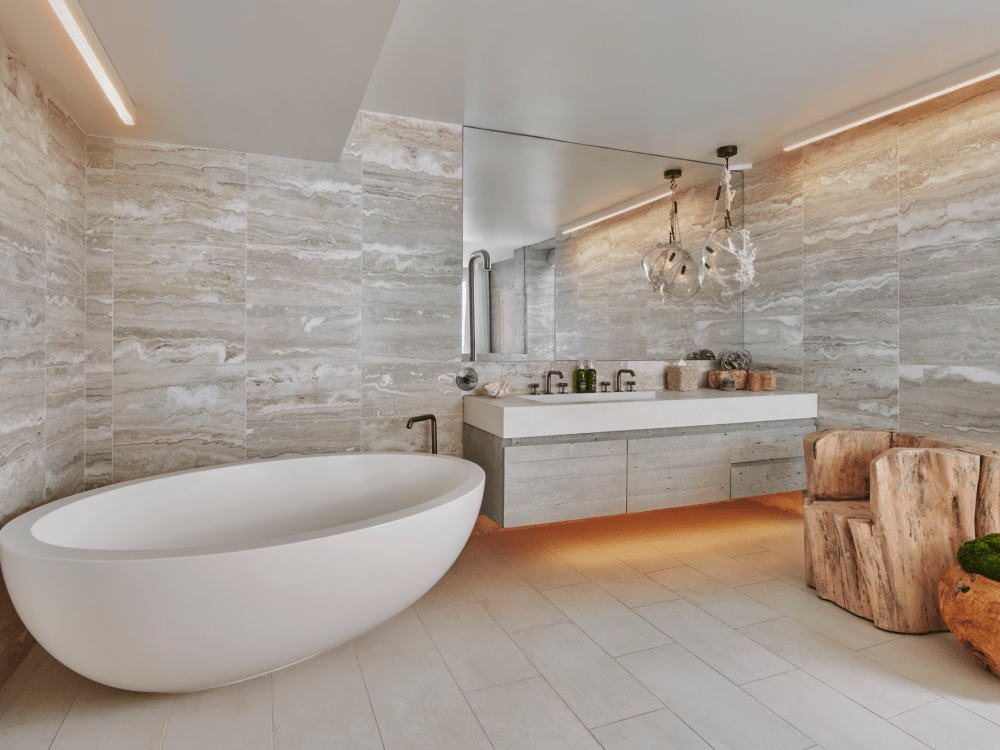 Modern bathroom with freestanding bathtub, dual sink vanity, and natural stone tiles. Features wooden chair and decorative hanging lights.