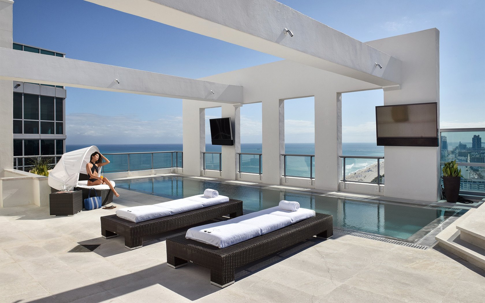 Rooftop pool area with ocean view, lounge chairs with white towels, woman sitting under canopy, and TV screens.