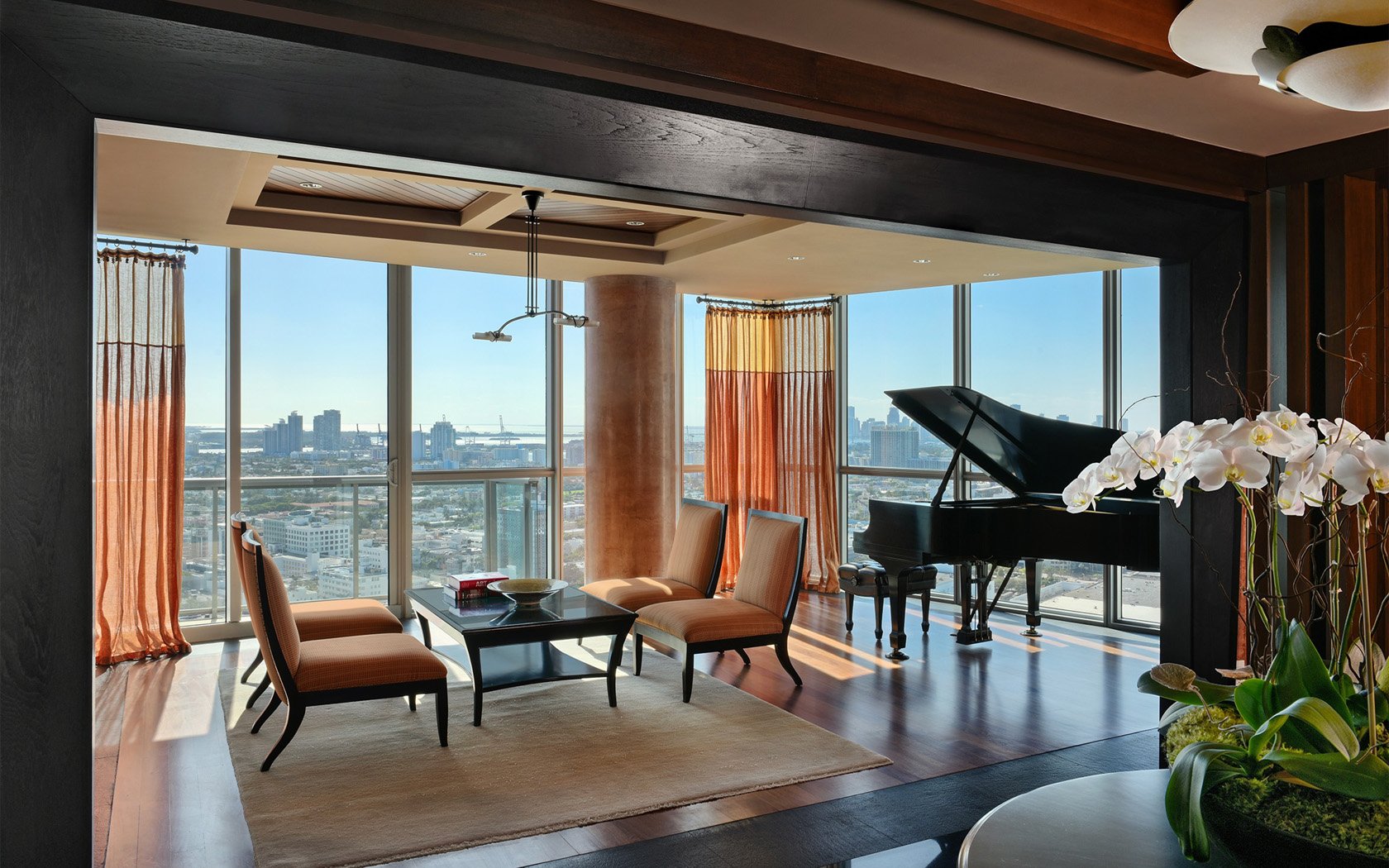 Luxury living room with panoramic city view, featuring a grand piano, modern chairs, and a coffee table on a rug, with potted white orchids in the foreground.