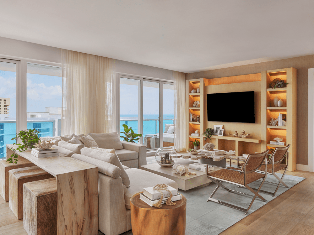 Modern living room with ocean view, featuring beige sofas, wooden stools, a flat-screen TV on a bookshelf wall, glass coffee table, and large windows with sheer curtains.